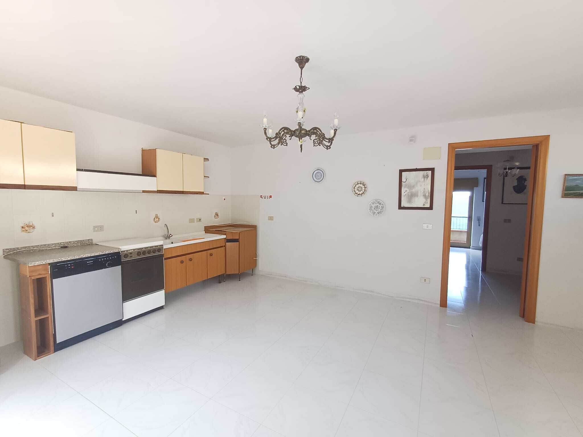 Spazio soggiorno cucina vuoto, cucina con mobili in legno, elettrodomestici e rivestimento semplice, pareti bianche e pavimento in piastrelle lucide in stile classico, dettagli decorativi come quadro e piatti, porta verso una stanza con balcone.