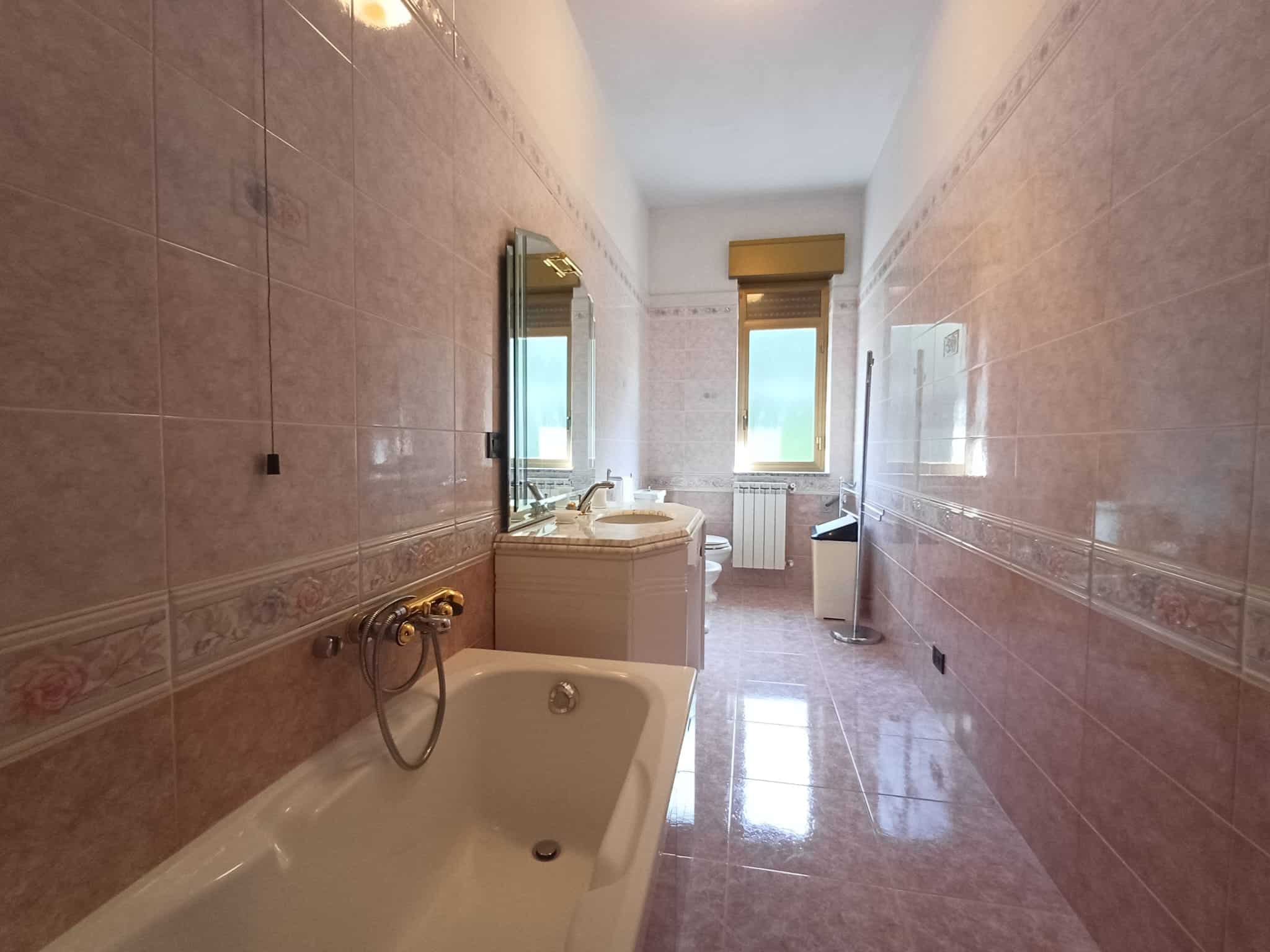 Bagno spazioso con piastrelle rosa e dettagli decorativi, finestra luminosa, lavabo con specchio, vasca e accessori moderni, ideale per case in vendita o affitto a Napoli.