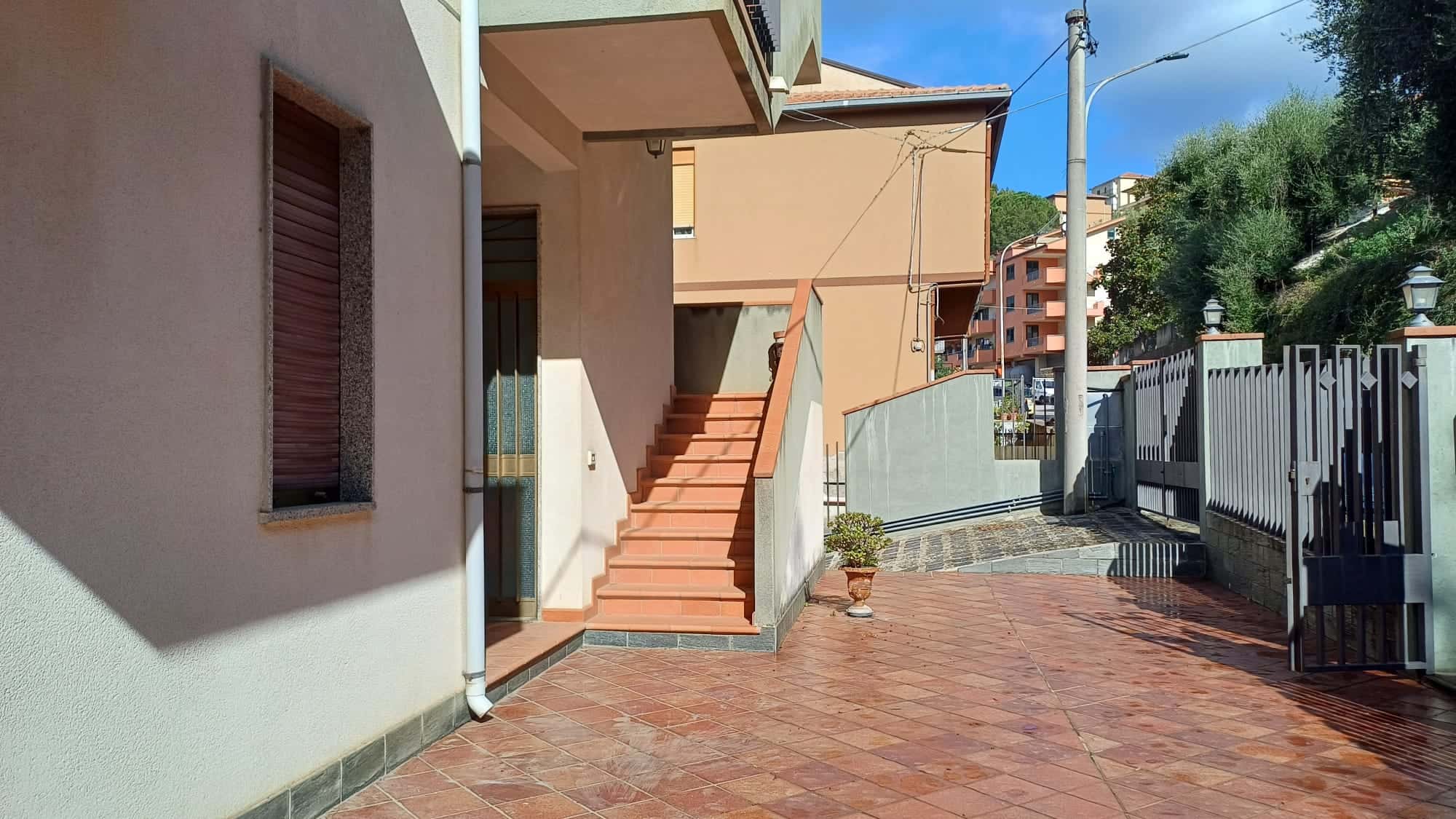 Scala esterna in mattoncini con ringhiera, ingresso di una casa con portafinestra e finestra, ambiente esterno ben curato, zona residenziale con edifici colorati sullo sfondo.