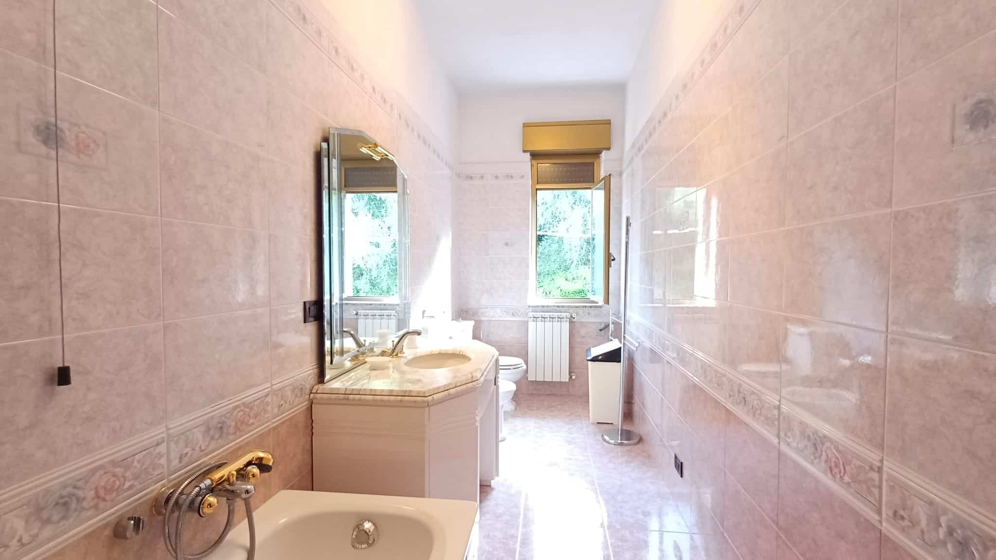 Stanza da bagno in stile classico con piastrelle rosa, lavabo con mobile in marmo, grande finestra con vista verde, radiatore e sanitari moderni. Ambiente luminoso e spazioso, ideale per l'arredamento di case di pregio.