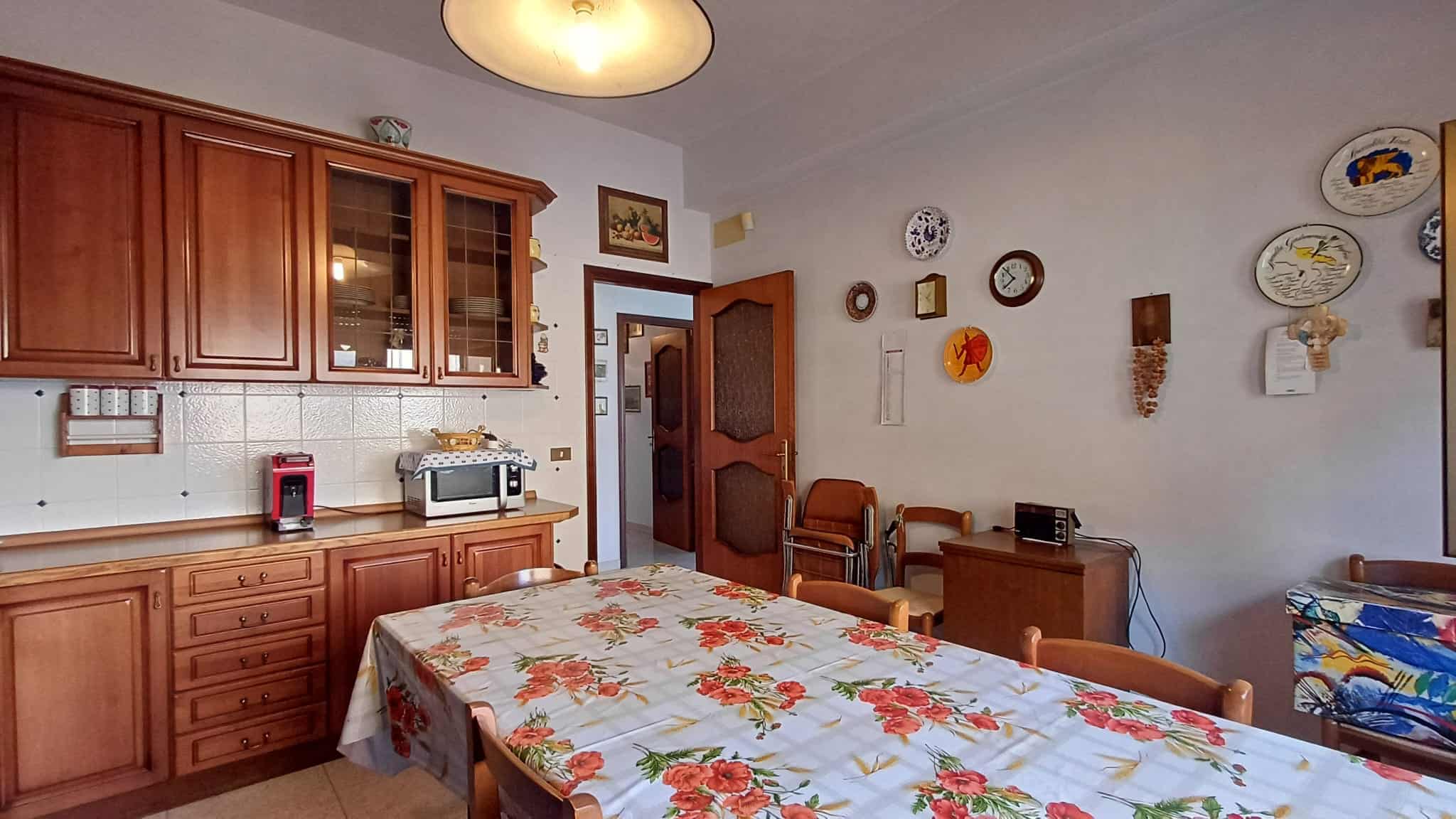 Elegante cucina con mobili in legno massiccio, tavolo con copertura floreale, elementi decorativi su parete, elettrodomestici funzionali, atmosfera calda e accogliente, ideale per vivere momenti conviviali.
