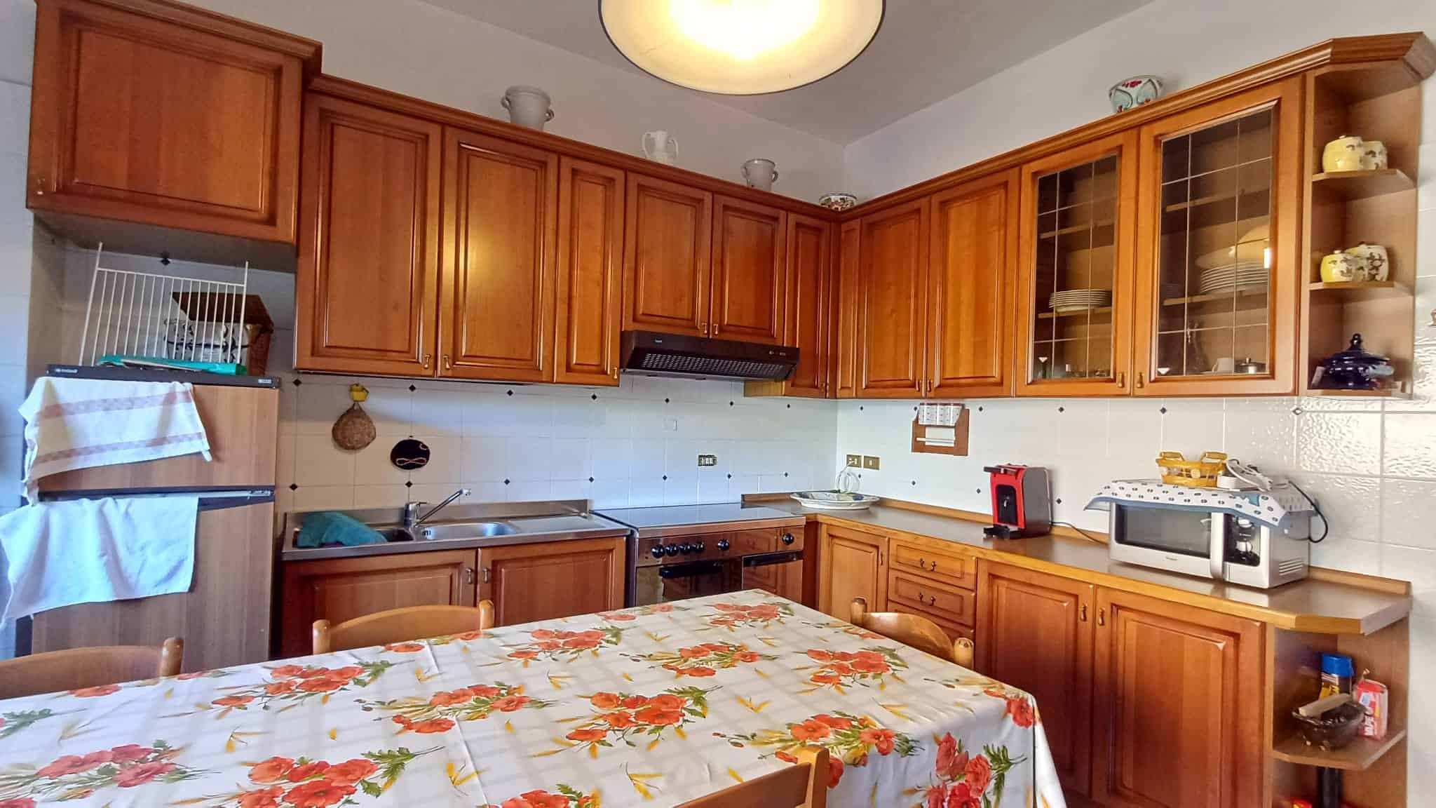 Fondo cucina in legno con pensili e ripiani, zona pranzo con tavolo decorato, elettrodomestici e utensili da cucina, ambiente caldo e accogliente.