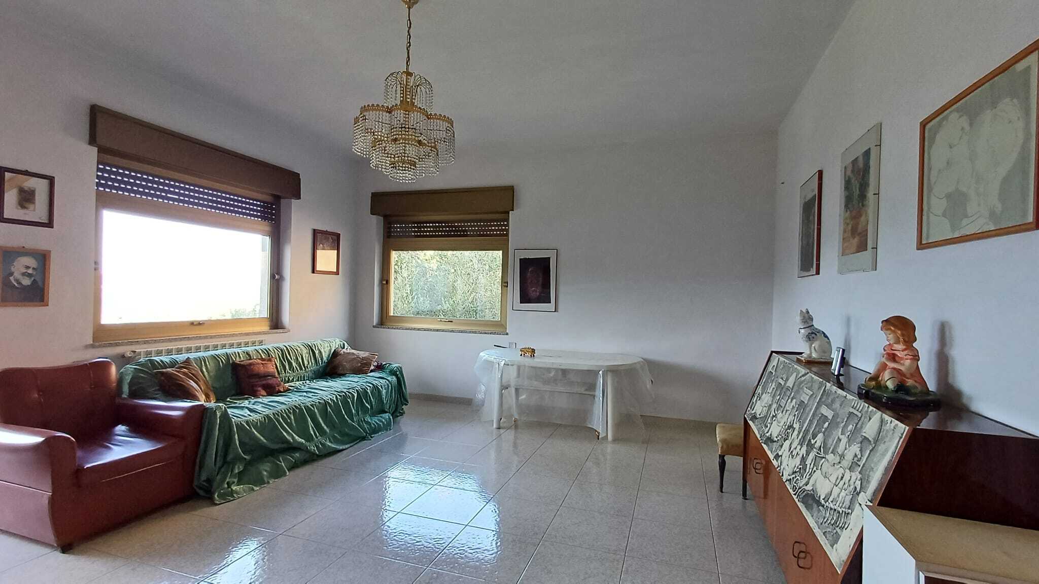 Soggiorno spazioso con divano verde, poltrona rossa e decorazioni artistiche sulla parete, luminoso grazie alle finestre e illuminato da lampadario retrò, ideale per vivere momenti di relax in casa.