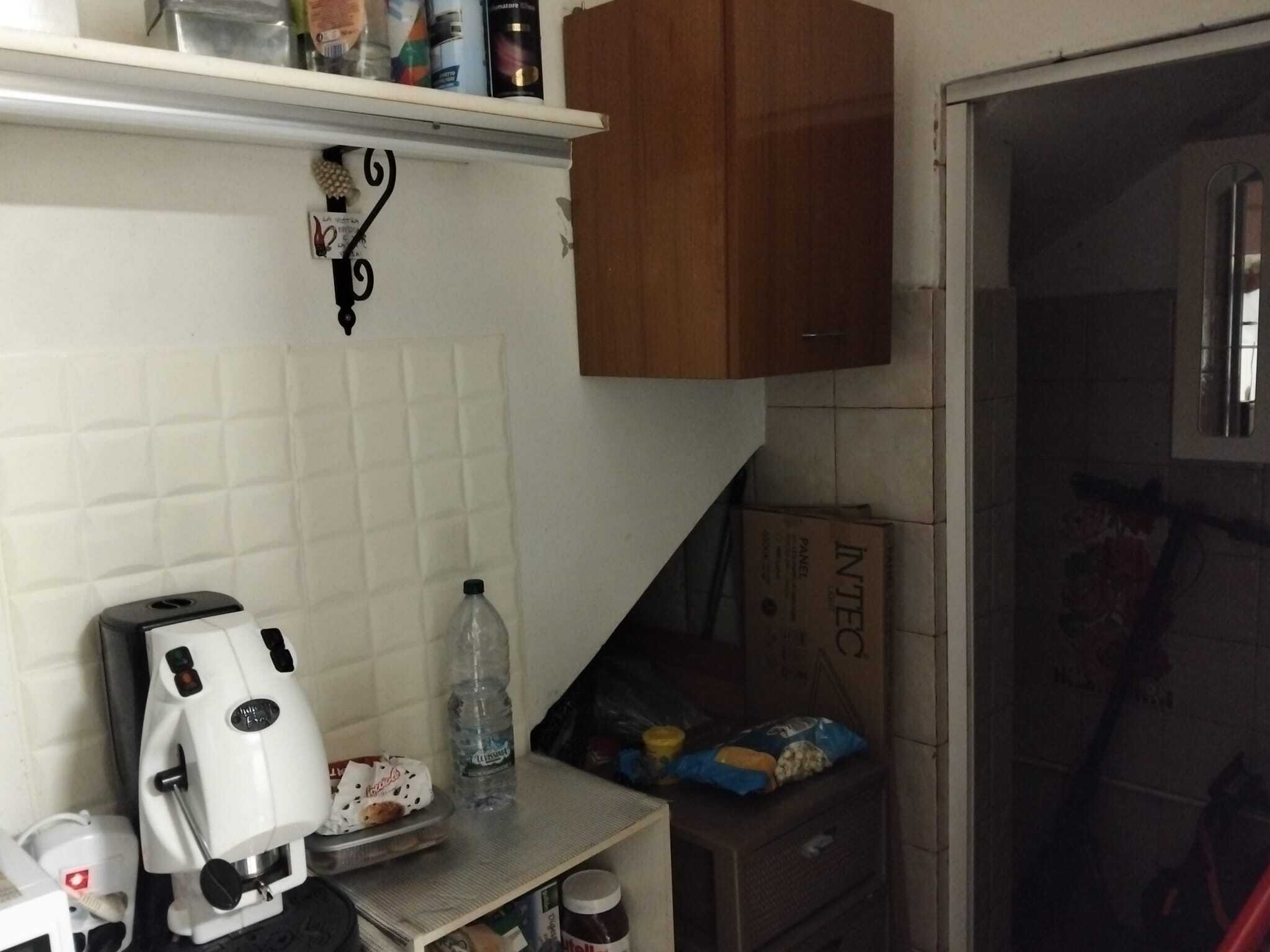 Pentola ad aria calda bianca, bottiglia d'acqua, ripiano con snack e utensili da cucina, armadietto in legno, stanza con ingresso in vetro e pareti di piastrelle beige, ambiente cucina abitabile.