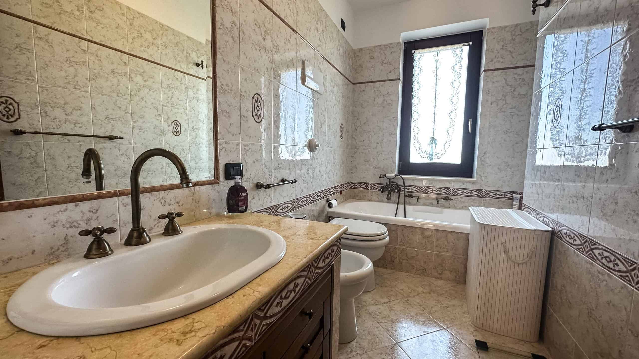 Lavabo in bagno con lavabo in ceramica e rubinetti in ottone, specchio grande, vasca da bagno con finestra, ripostiglio in plastica, interni di alta qualità in stile classico.