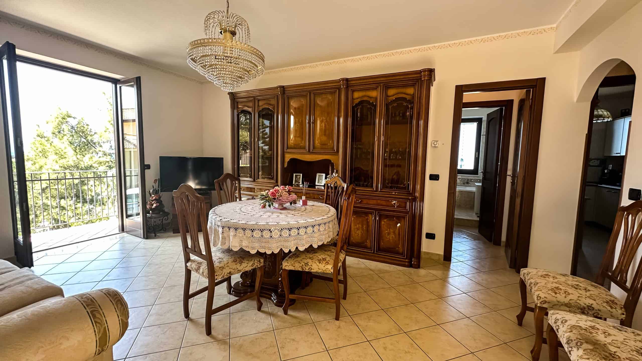 Ampio soggiorno con porta finestra che dà sul balcone, arredi vintage, grande mobile in legno su parete, tavolo rotondo con tovaglia e fiori, televisore, pavimenti in ceramica, ambiente accogliente per la casa.