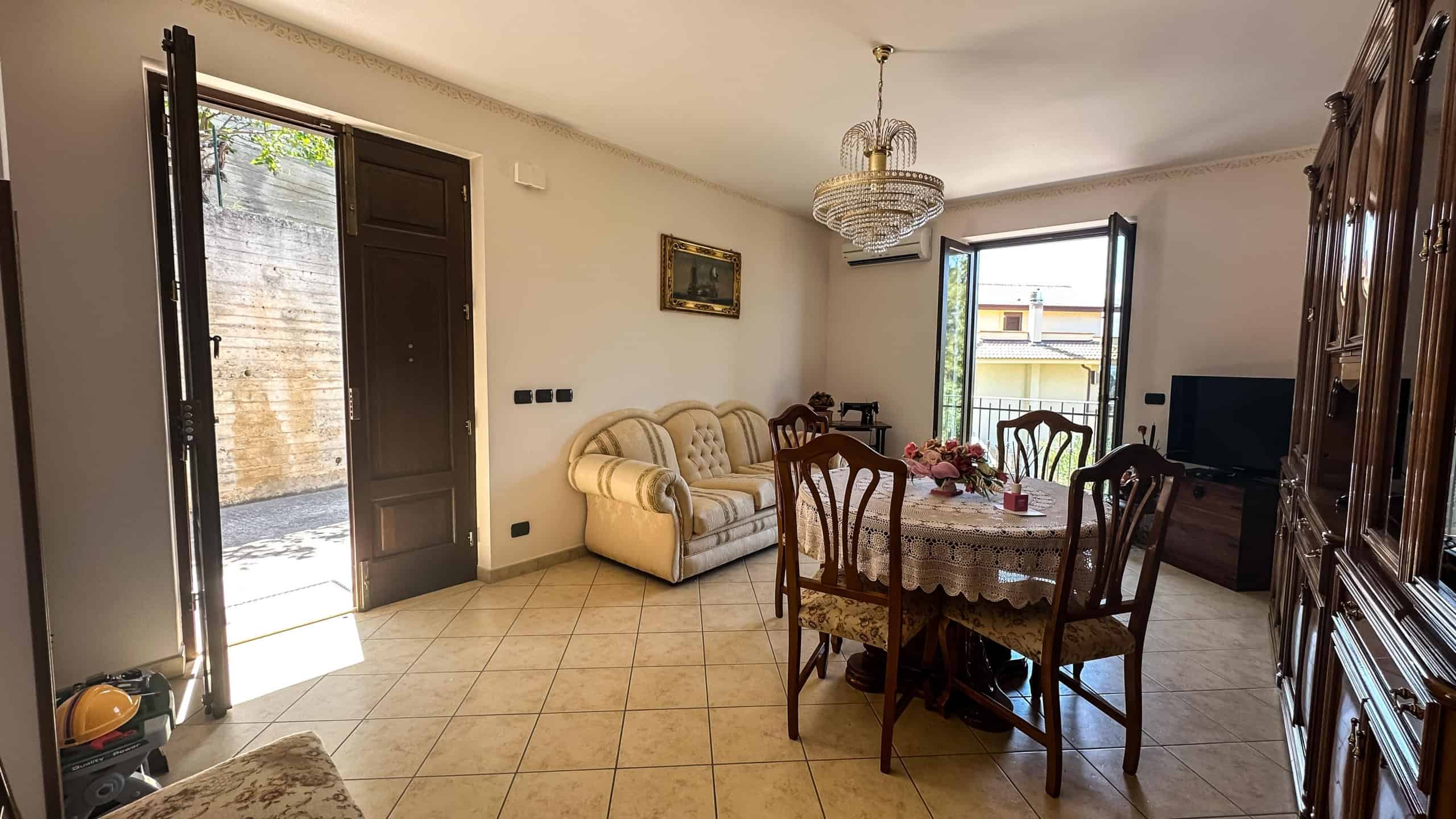 Vivace soggiorno con pareti bianche, divano in stile classico e tavolo da pranzo con tovaglia ricamata, illuminato da cristallino lampadario e illuminazione naturale da finestre e porta del balcone.