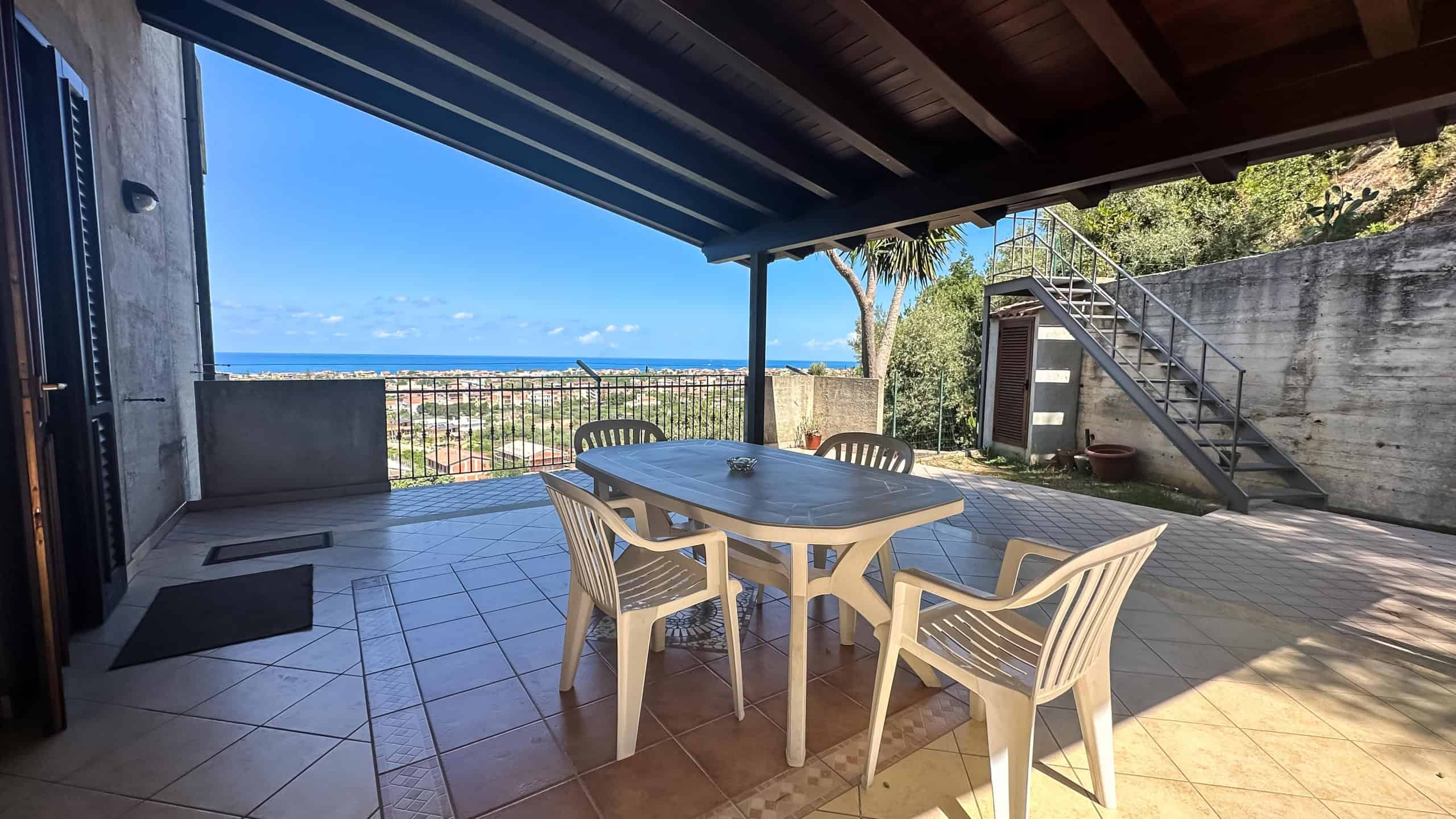 Vista mare dalla terrazza con tavolo e sedie in plastica, ambiente esterno soleggiato, con scale in metallo e pareti in cemento, ideale per relax e convivialità.