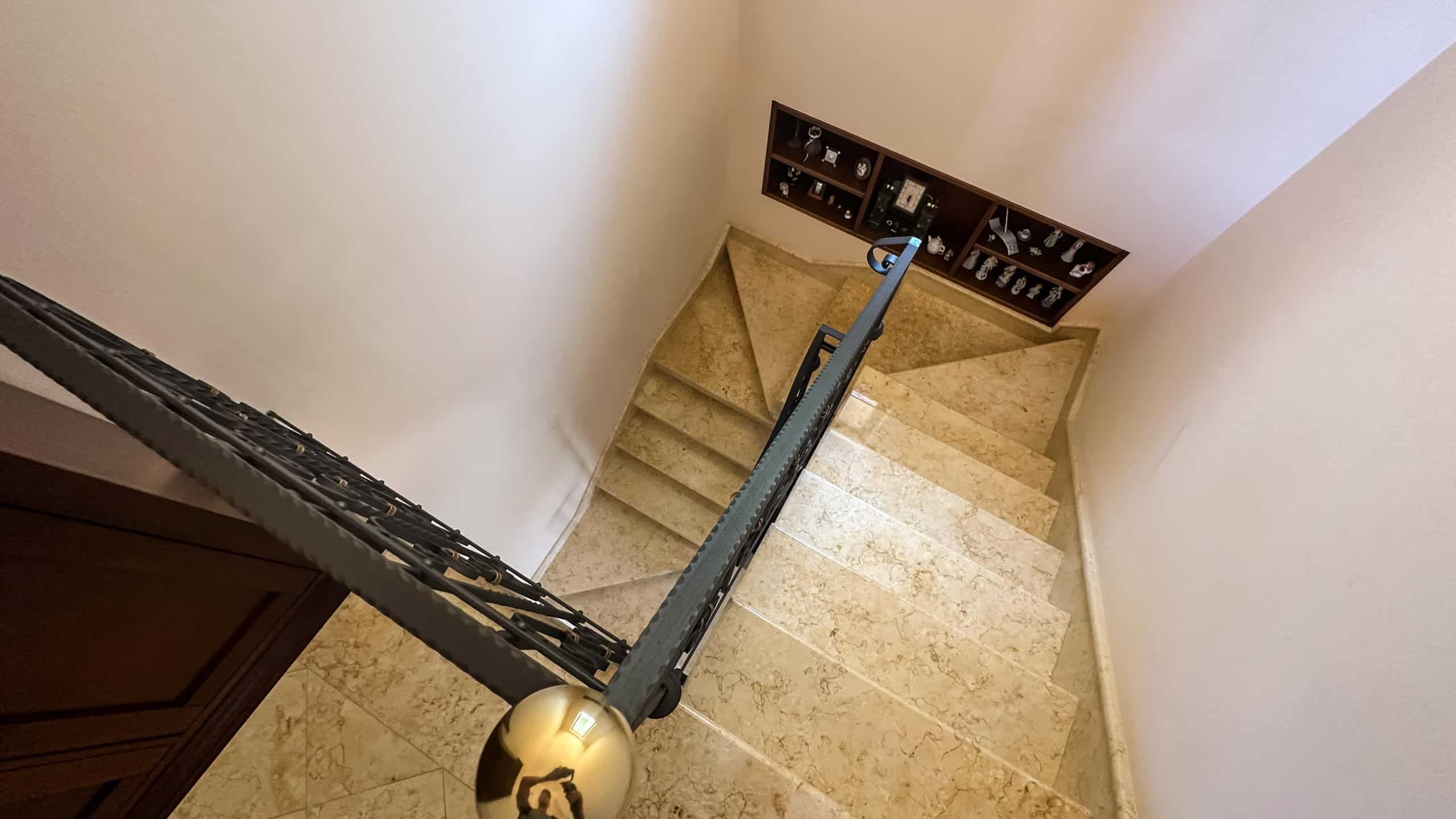 Scale internamente in marmo beige con ringhiera in ferro battuto, vista dall’alto di una scala che conduce a un piano superiore con mensole decorative e oggetti d’arredo eleganti, ambiente raffinato e di design.