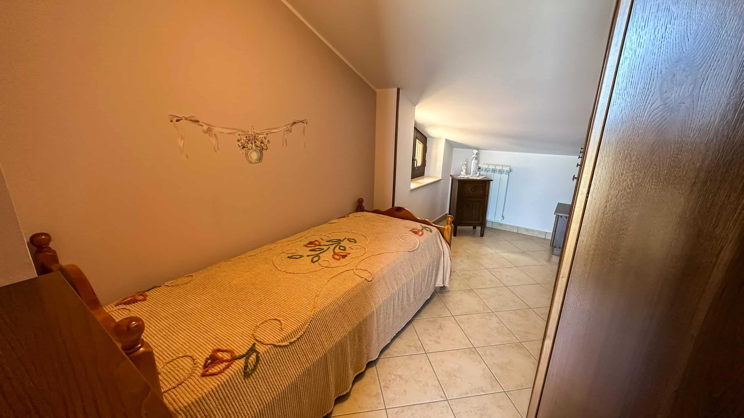 Letto singolo con coperta decorativa in una camera mansardata accogliente e luminosa, con casseruola in legno e decorazioni floreali, ideale per affitti di breve e lungo termine.