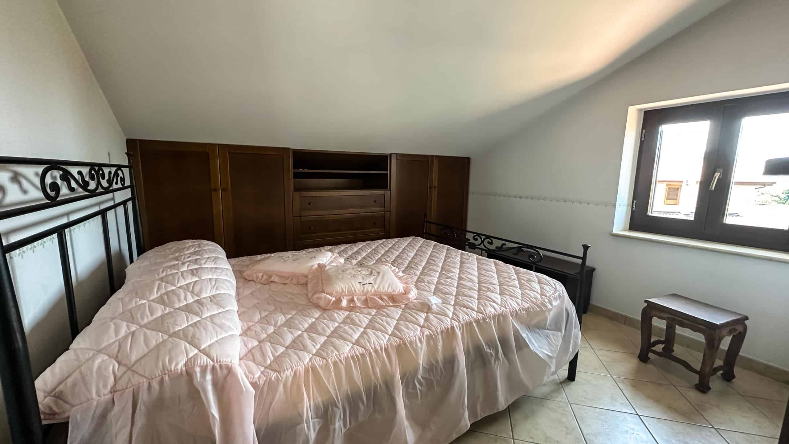 Letto matrimoniale in una camera spaziosa con pareti bianche, finestra panoramica e arredi in stile classico, perfetto per affitto case vacanze o immobili residenziali, ambiente luminoso e accogliente.