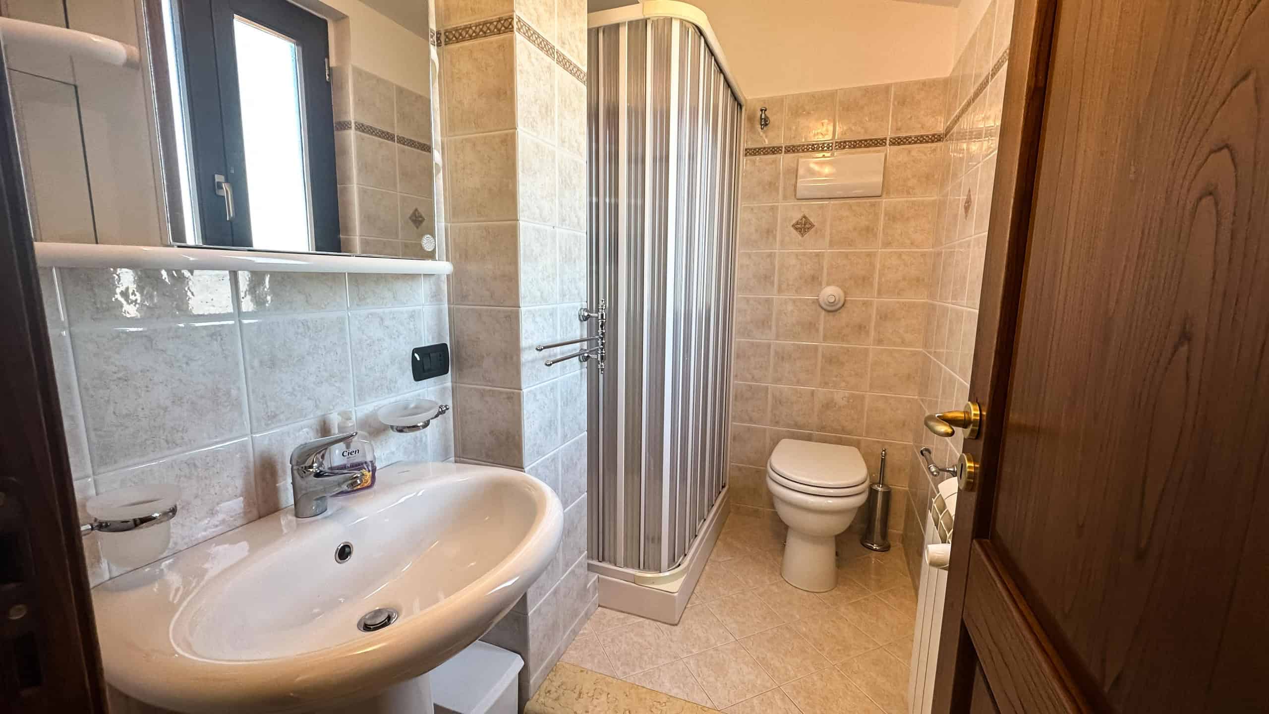Accogliente bagno con lavabo, doccia e WC in stile classico, rivestito con piastrelle beige e dettagli decorativi, ideale per case e appartamenti moderni o tradizionali.