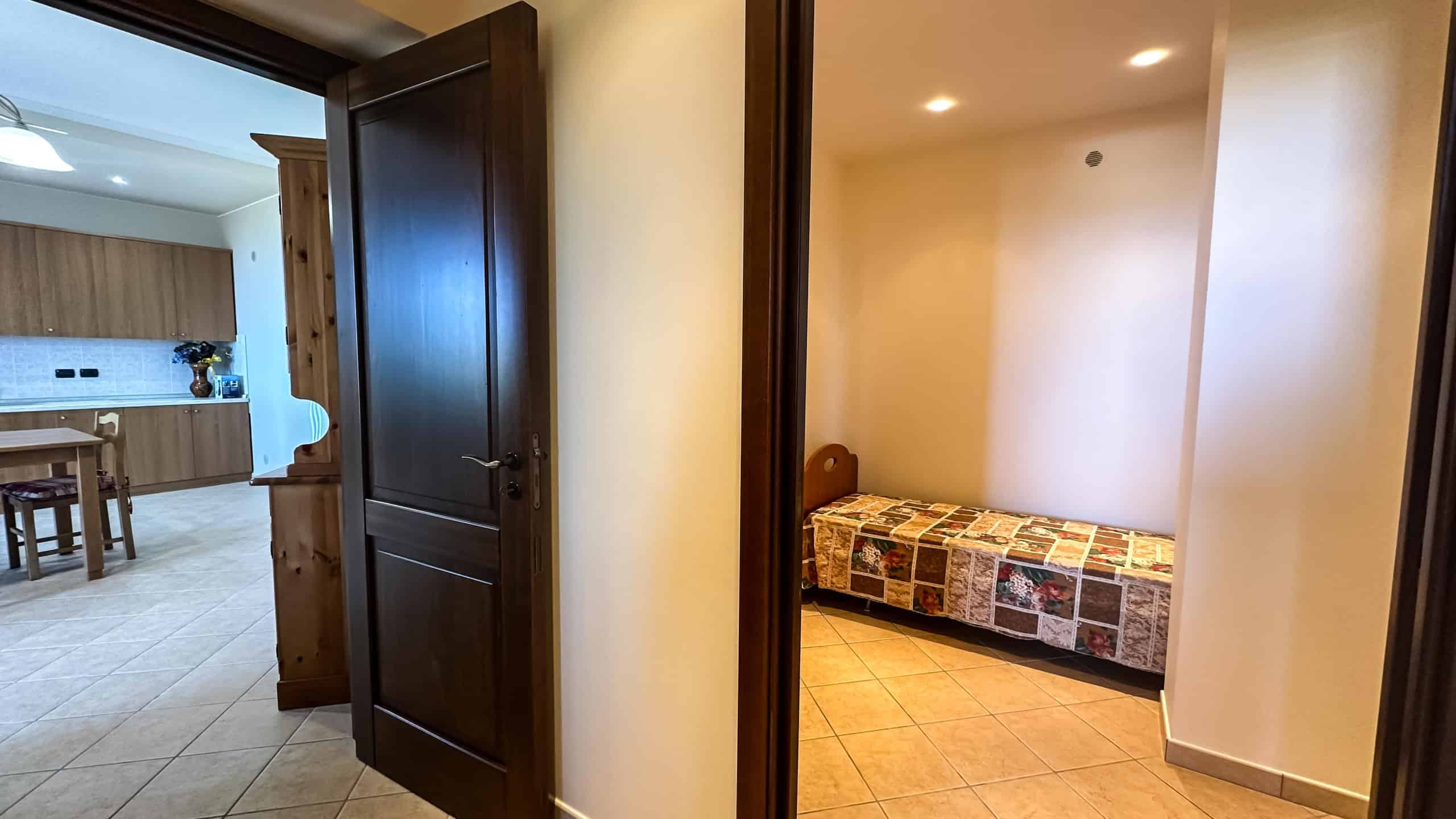 Camera con porta in legno che si apre su un soggiorno e una camera da letto con letto singolo e mobili in stile classico, interni di appartamento in affitto o vendita.