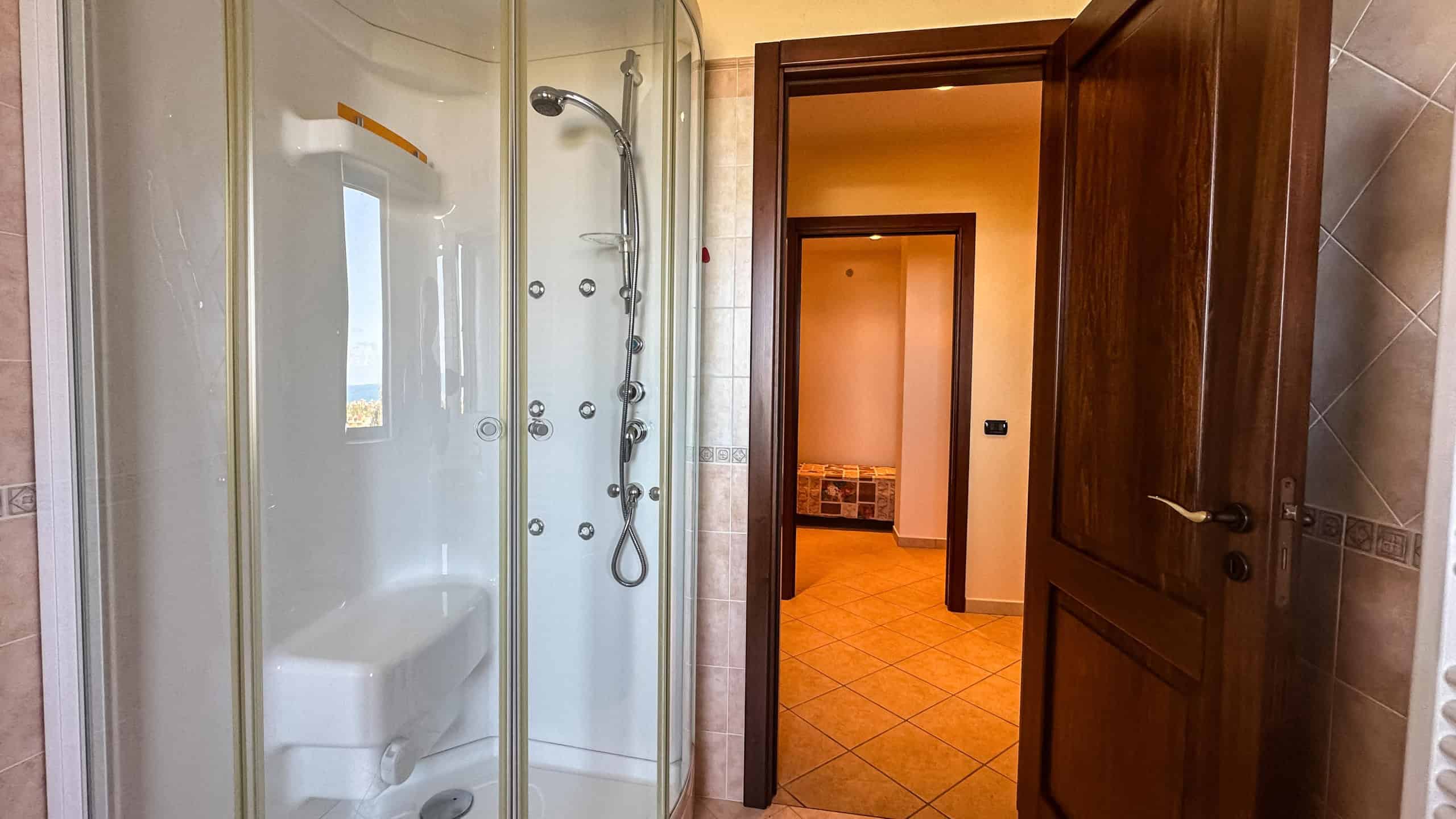 Ripresa di un bagno con doccia idromassaggio, tenda in vetro, pareti in ceramica beige e porte in legno scuro, vista su corridoio con pavimentazione in piastrelle arancioni, ambiente elegante e funzionale.