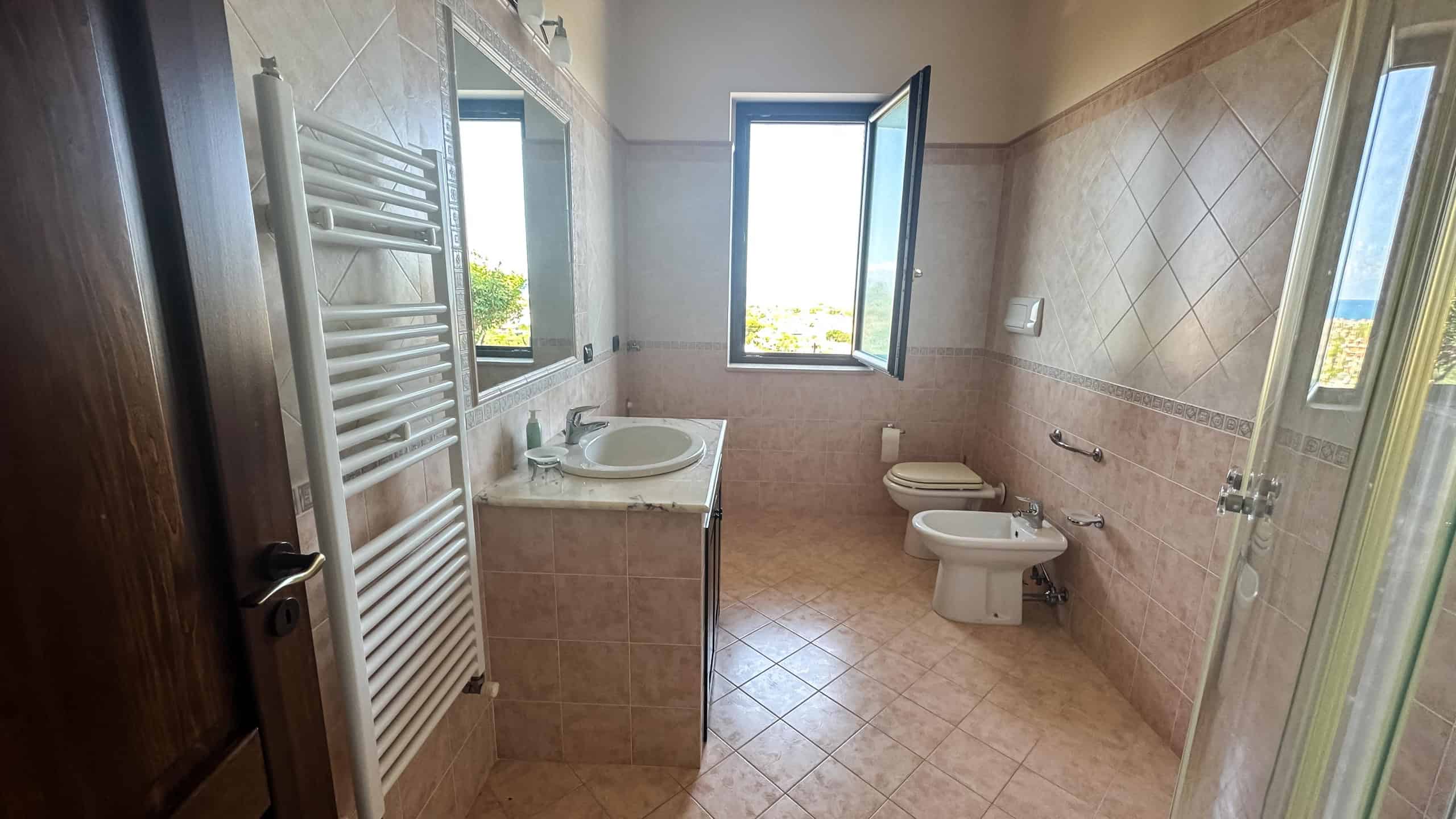 Ampia stanza da bagno con finestra, lavabo con piano in marmo, WC e bidet, riscaldamento a muro, pareti e pavimenti rivestiti in piastrelle beige, ambiente luminoso e arioso.