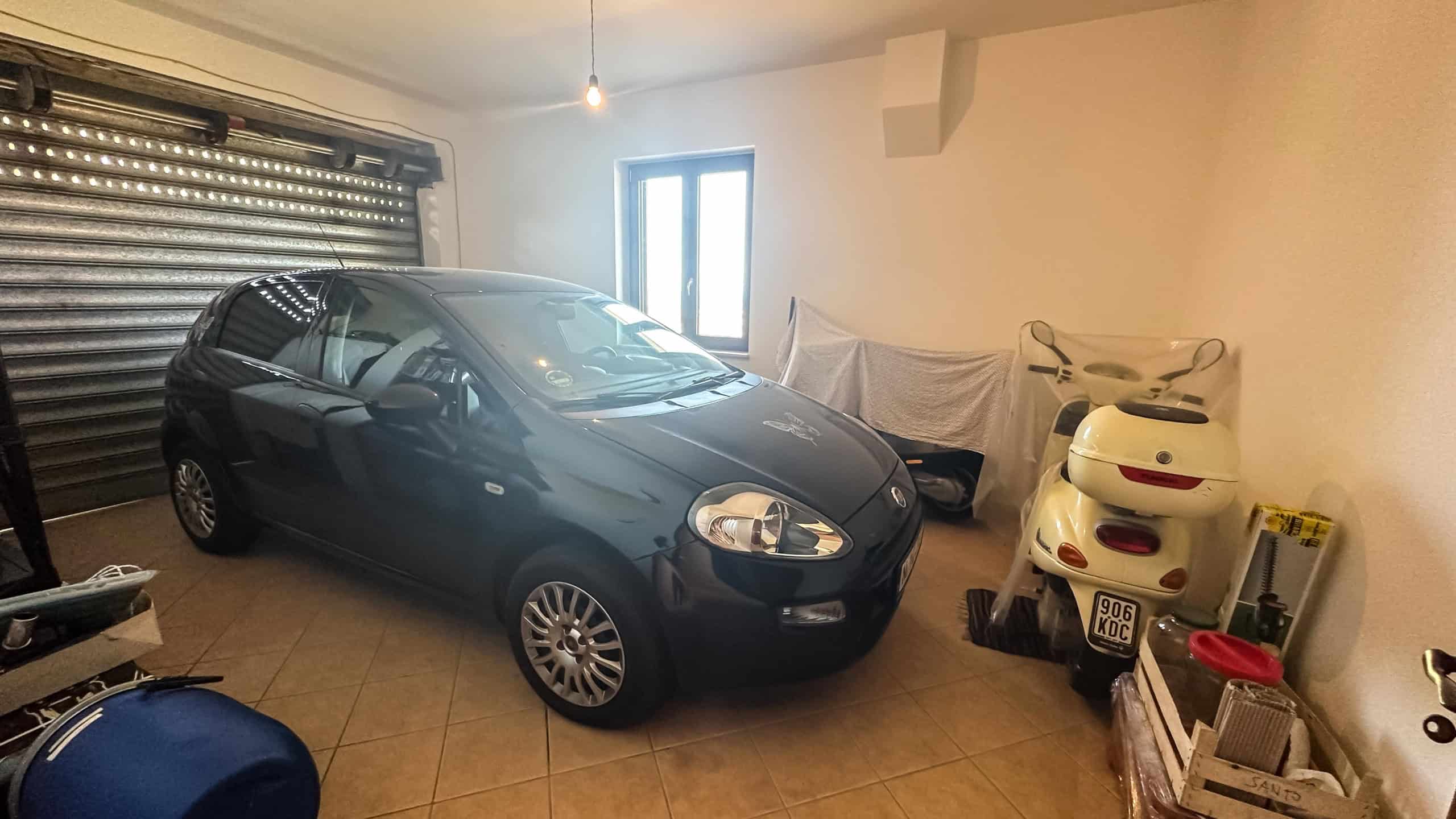 Auto compatta nera in garage con pareti bianche e finestra, spazio di deposito per moto e attrezzi, ambiente ben illuminato, Fiat Punto, piani in ceramica beige, organizzazione efficiente degli spazi.