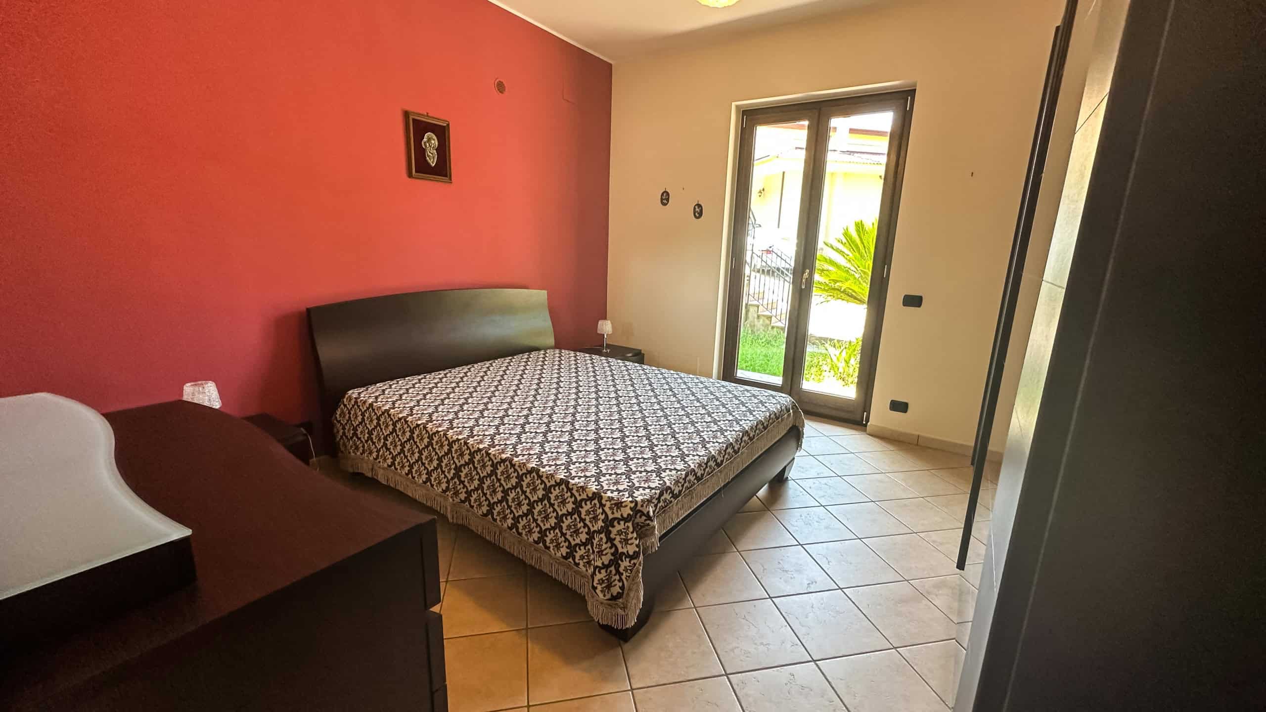 Letto matrimoniale con testiera in legno naturale e letto singolo, spazio notte luminoso con accesso a giardino privato, arredamento moderno, elegante e funzionale, ideale per affitti turistici o residence.