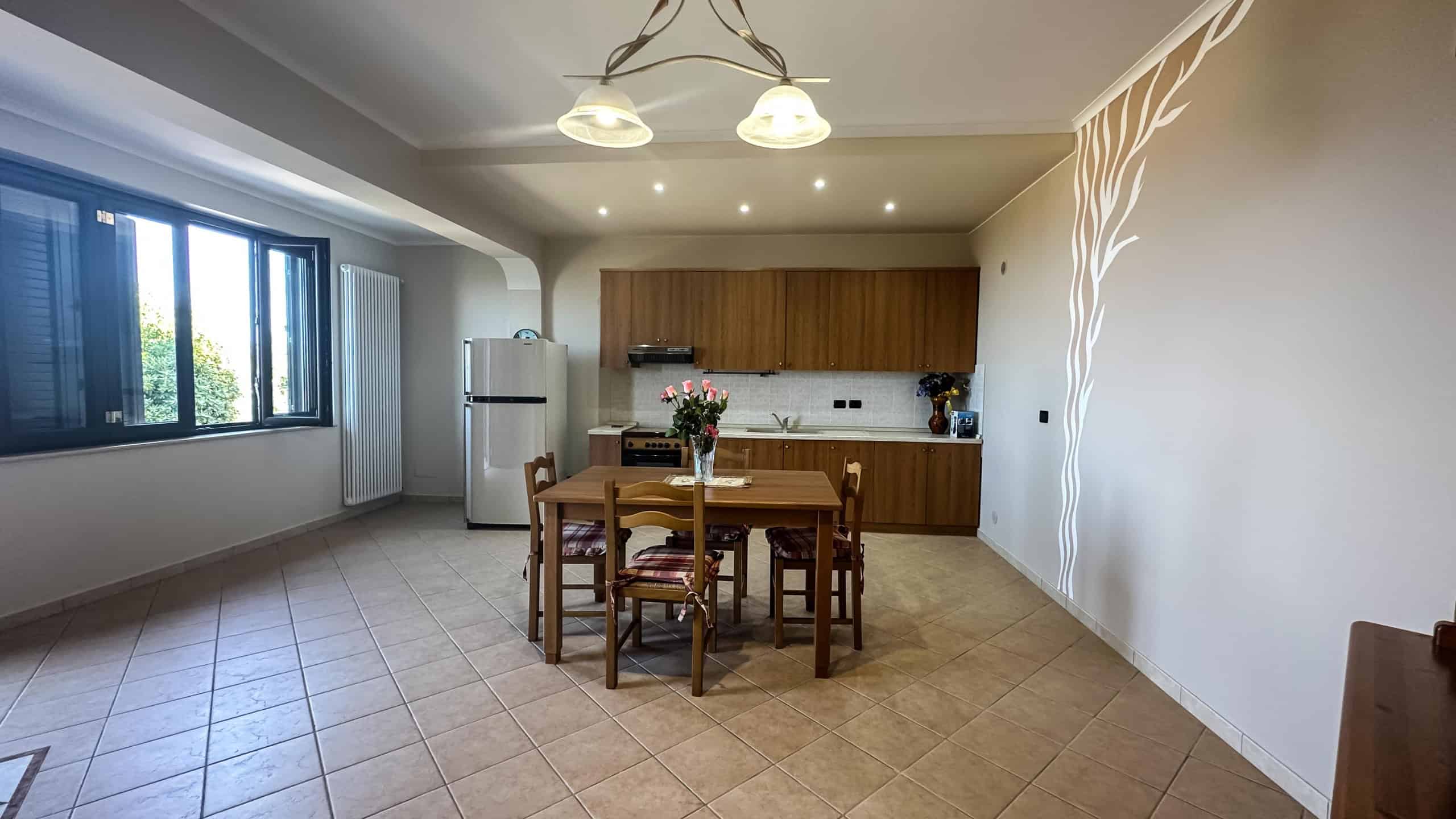 Vasta cucina soggiorno con zona pranzo, pareti bianche, mobili in legno, grande finestra con vista verde, decorazioni moderne, elettrodomestici inclusi, atmosfera calda e accogliente.