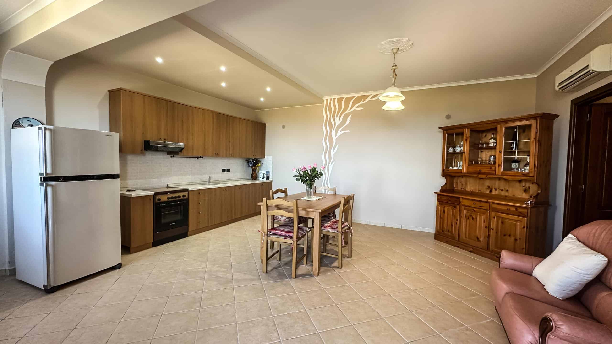Spazio soggiorno e cucina in stile tradizionale con mobili in legno, zona pranzo con bouquet di fiori, pavimento in ceramica, parete decorata con motivo tree, e aria condizionata.