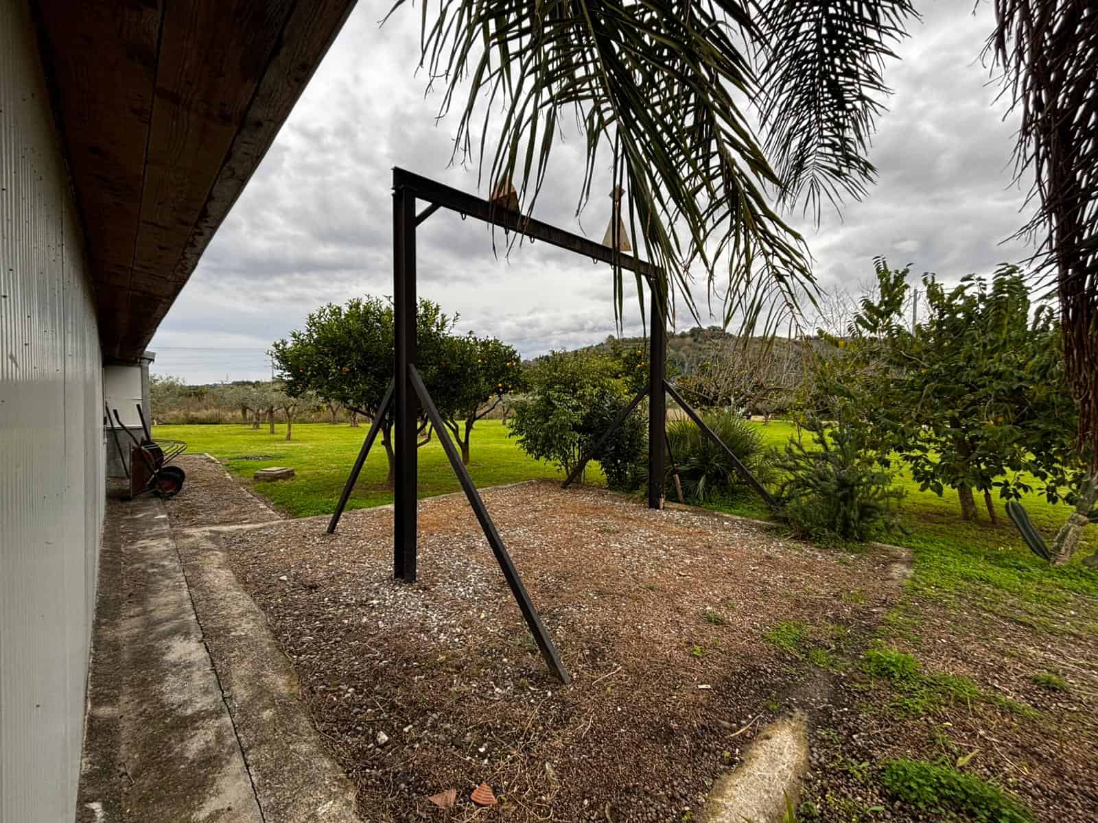 Alt: Struttura per altalena in giardino con alberi e vegetazione verde sotto cielo nuvoloso.