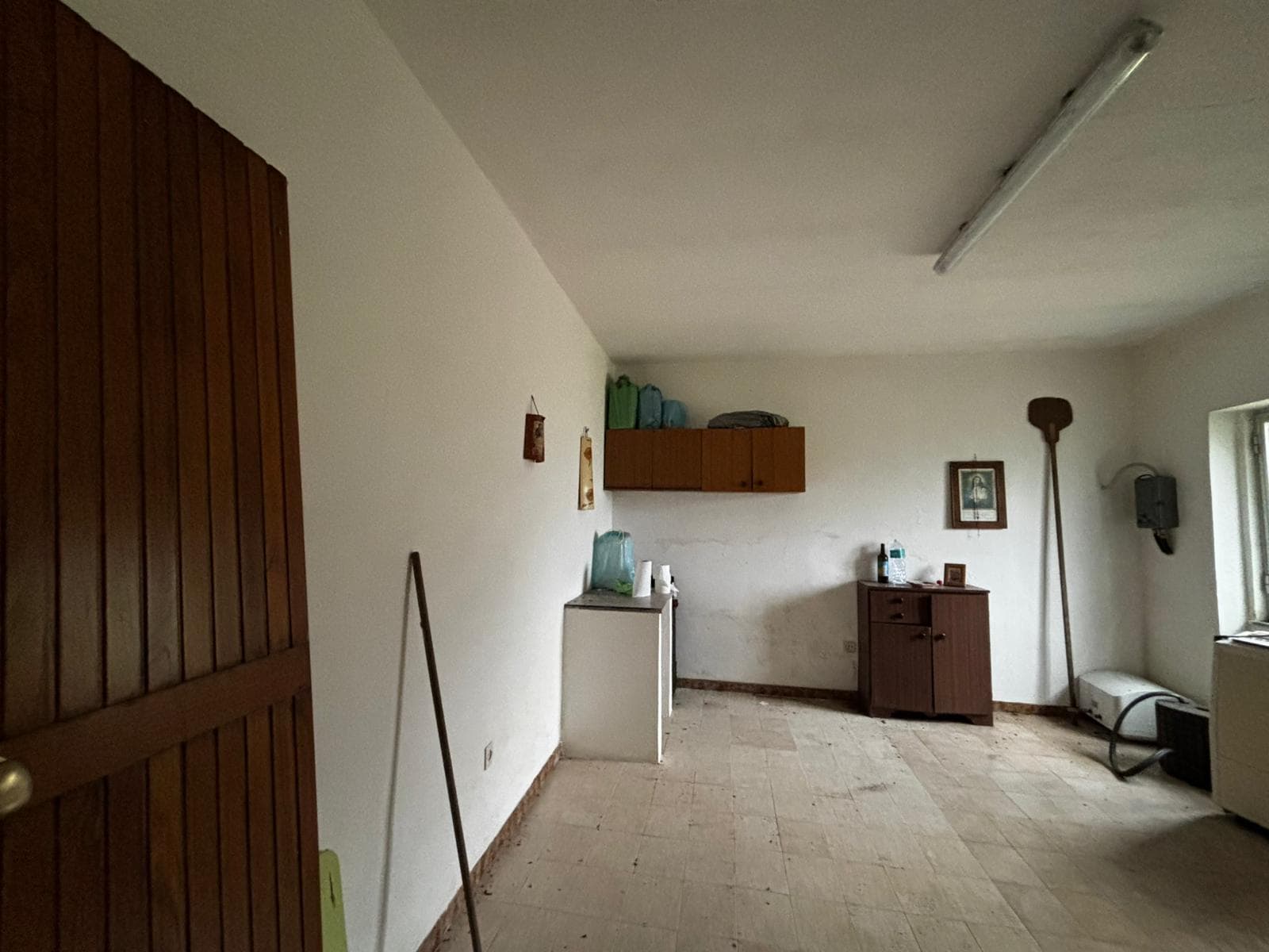 1. Interno di una stanza vuota e trascurata con pareti bianche, pavimento in legno usurato e pochi mobili di epoca e stile semplice a NextCasa.