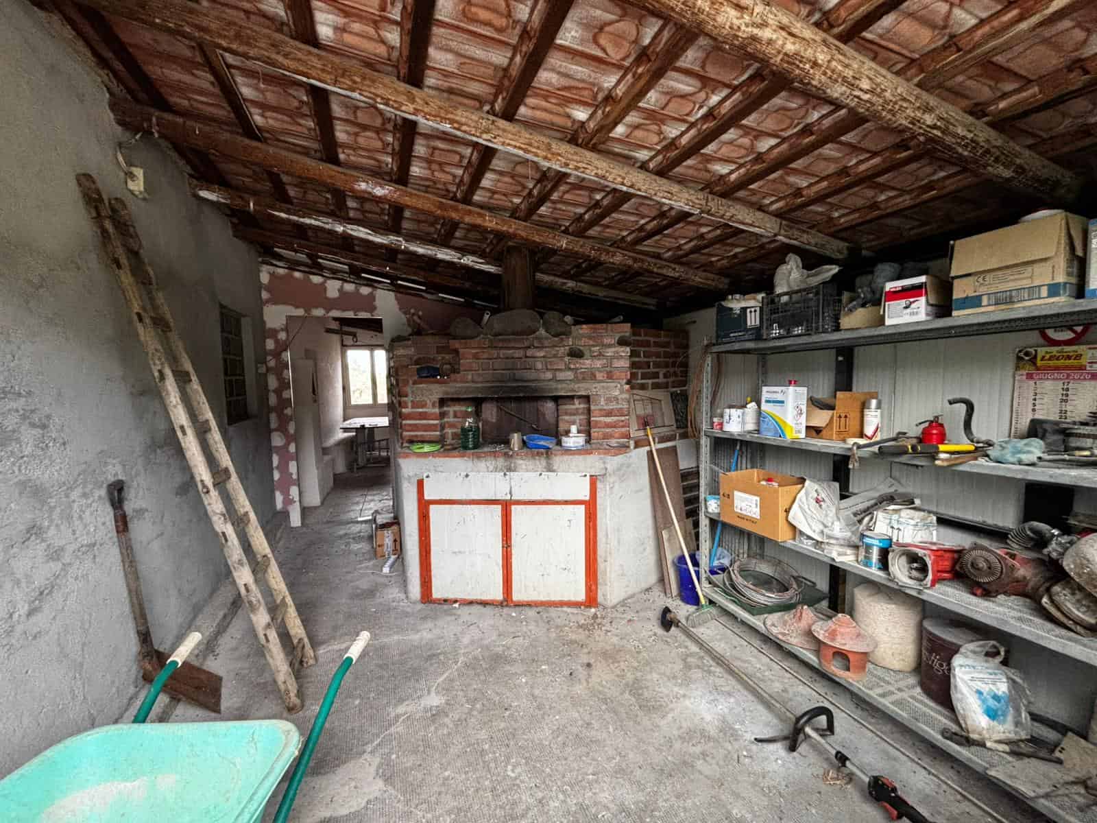 Ripostiglio in sottotetto con scaffali pieni di attrezzi e materiali da bricolage, muro in cemento, pareti non finite e forti travi di legno sul soffitto, ambiente rustico e funzionale.