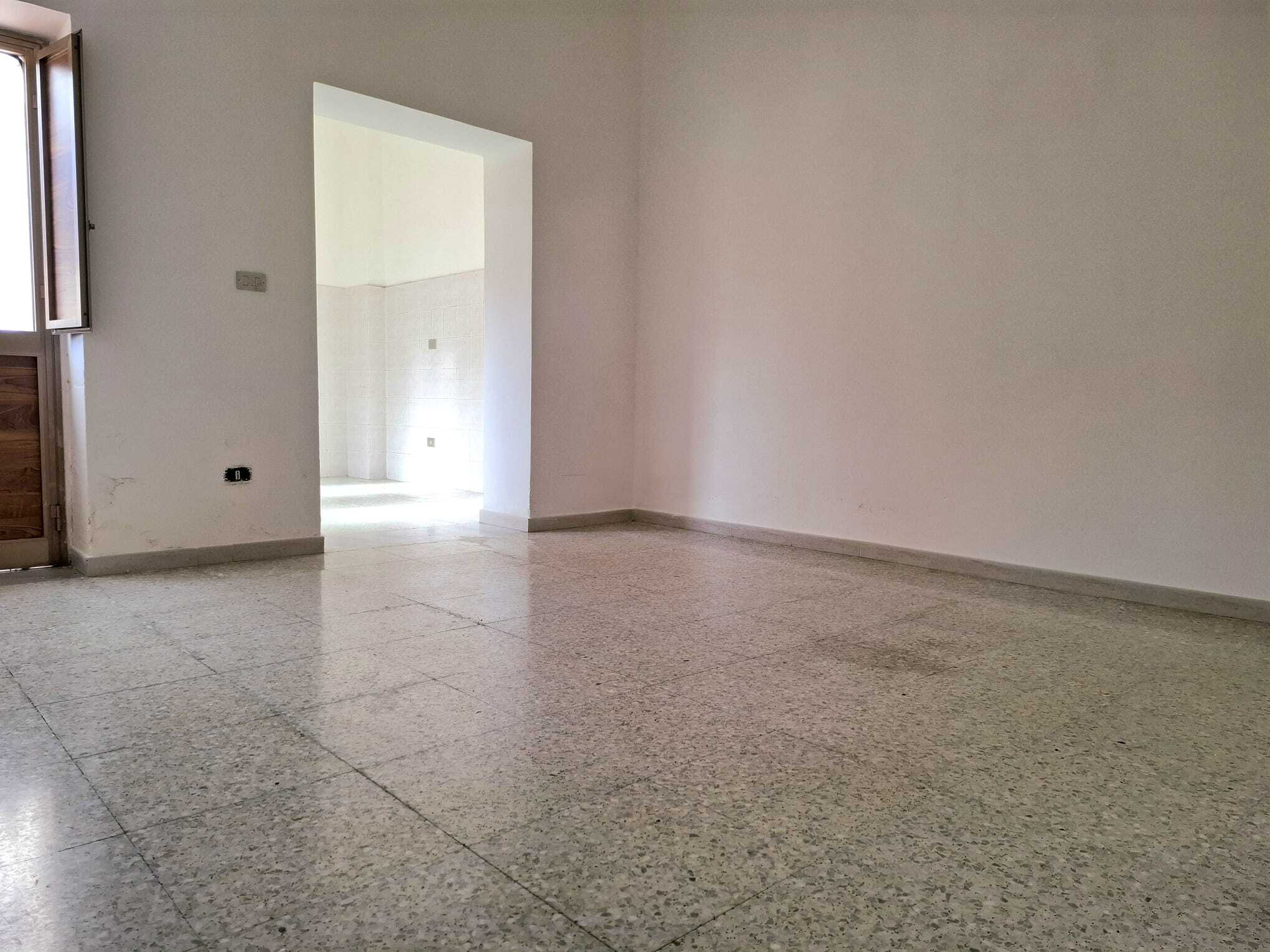 Ampia stanza vuota con pareti bianche e pavimento in piastrelle, ideale per ristrutturazione o arredamento personalizzato. Spazio luminoso e versatile, perfetto per abitazione o ufficio.
