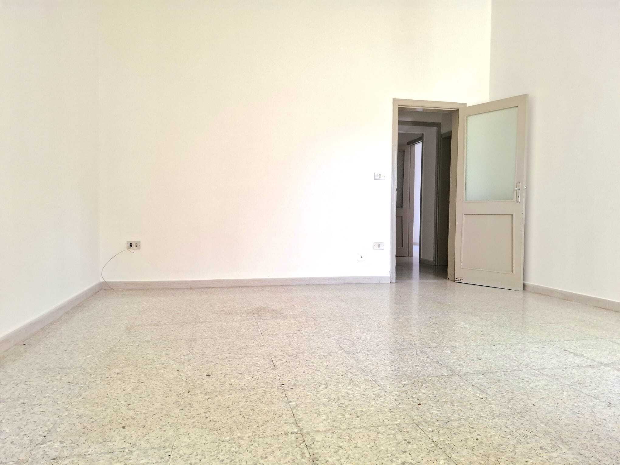 Ampio soggiorno vuoto con pareti bianche e pavimento in piastrelle, porta aperta che conduce a un'altra stanza, ambiente luminoso e spazioso ideale per arredamento personalizzato.