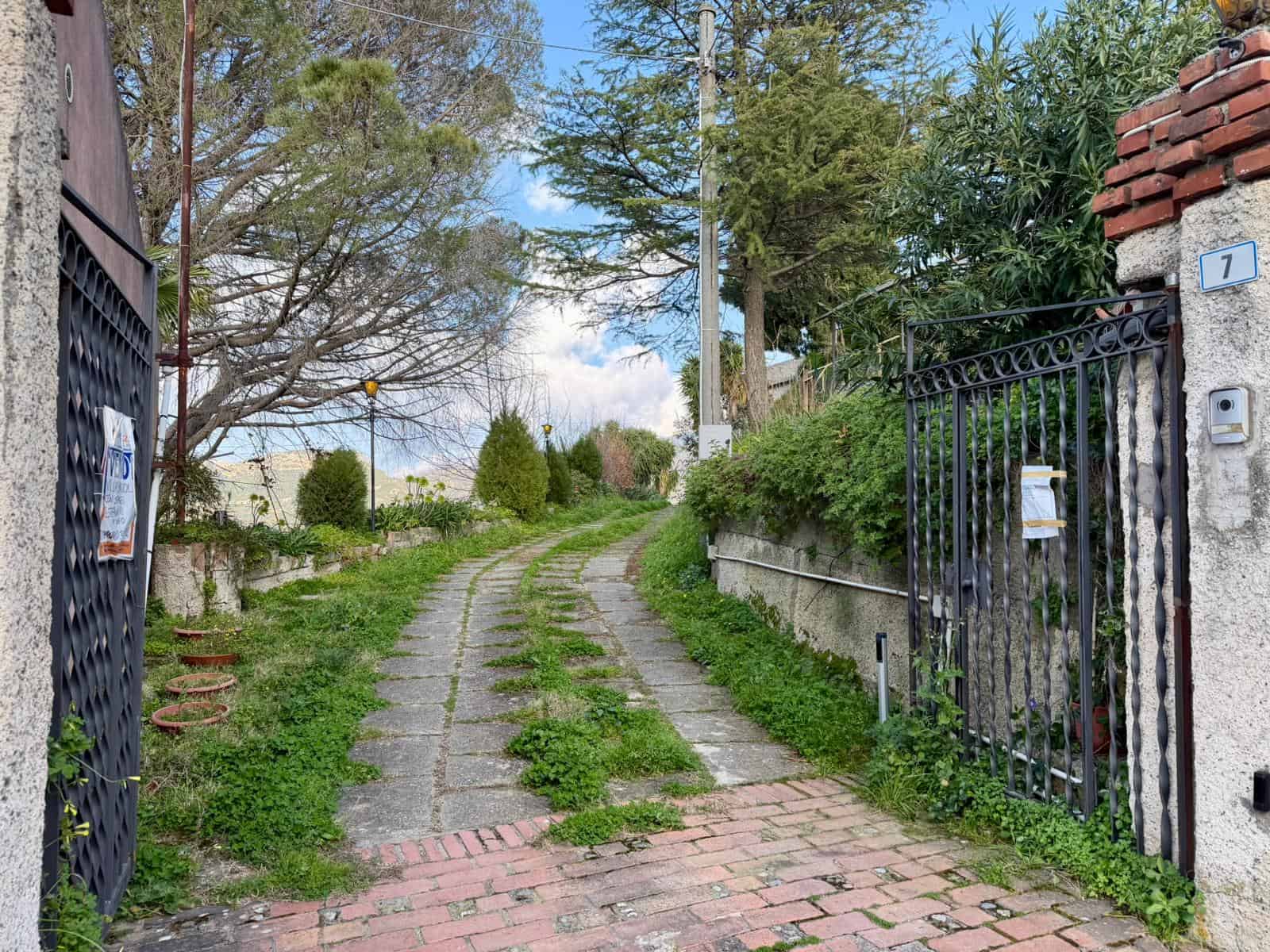 Accesso privato con strada sterrata e cancello in ferro, circondato da vegetazione e alberi, in zona residenziale tranquilla.