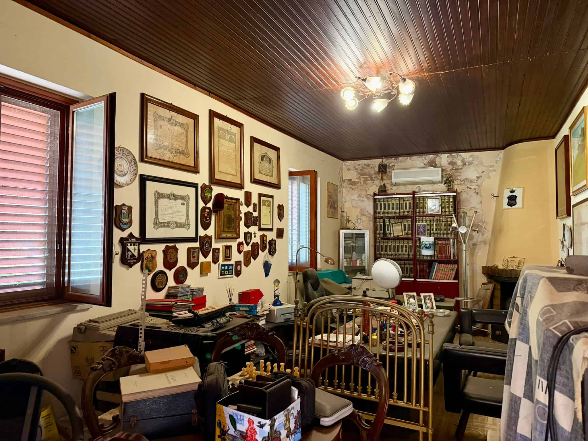 Interno di un soggiorno accogliente con parete decorata, mobili vintage e libri, illuminato da luce naturale e lampada da terra, stile classico e atmosfera calda.