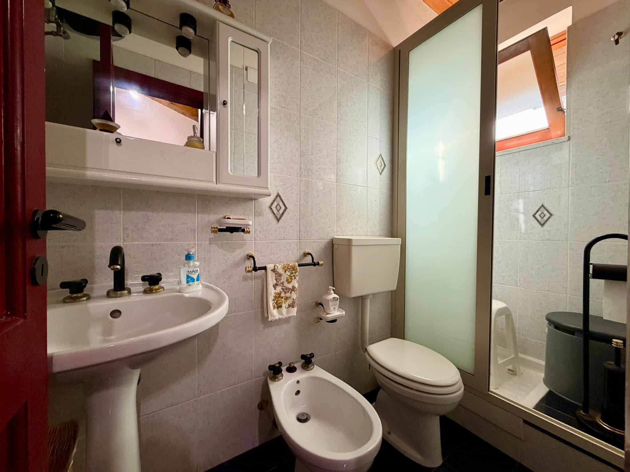 Lavabo e servizi igienici moderni in bagno con pareti in piastrelle chiare, doccia separata con porta in vetro e finestra per ventilazione naturale, ideale per affitti a breve termine o residenze di c.