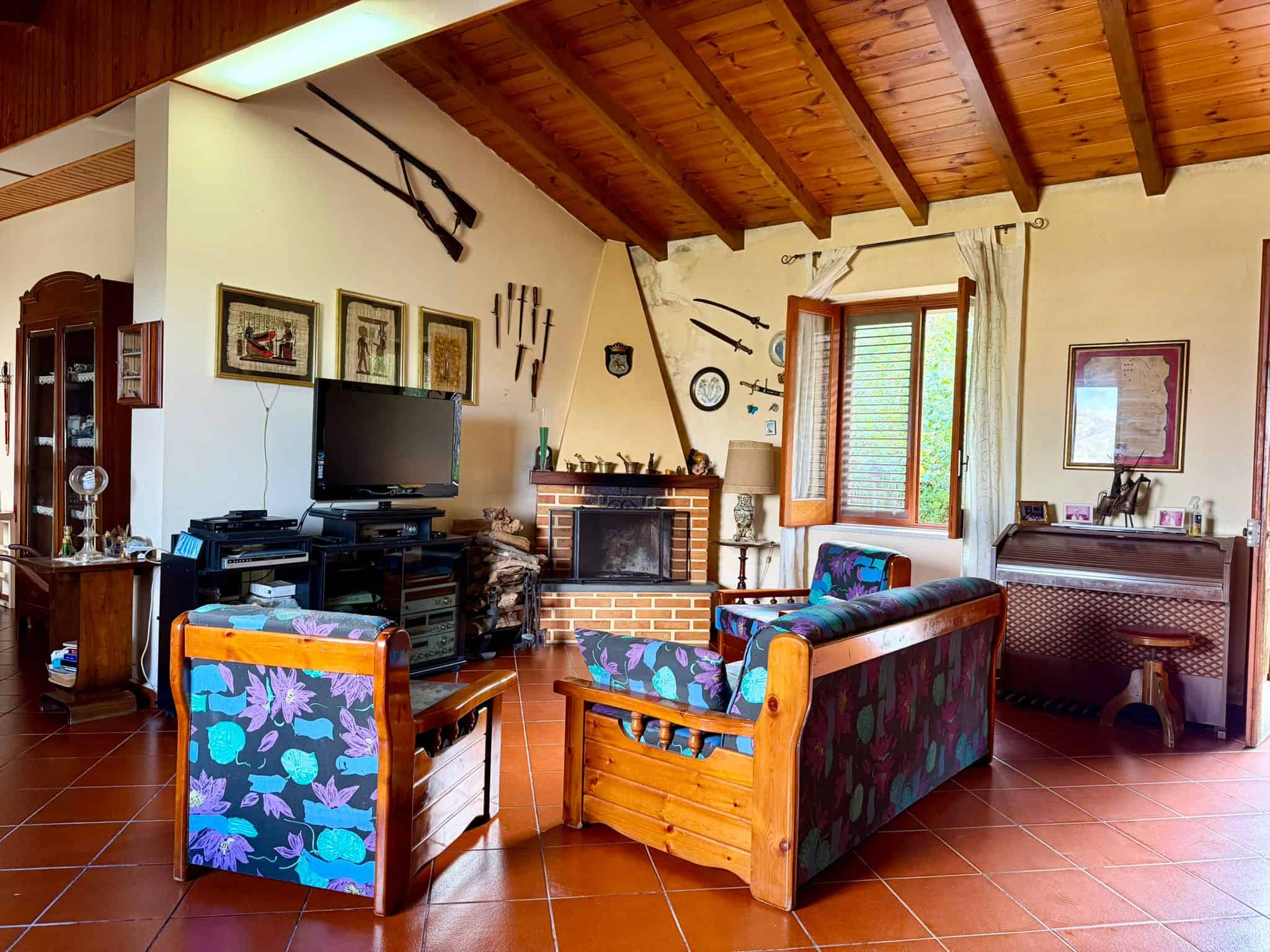 Interno di soggiorno rustico con mobili in legno, camino, pareti decorate con armi antiche e oggetti d'epoca, grande finestra con vista sul giardino, pavimento in terracotta.