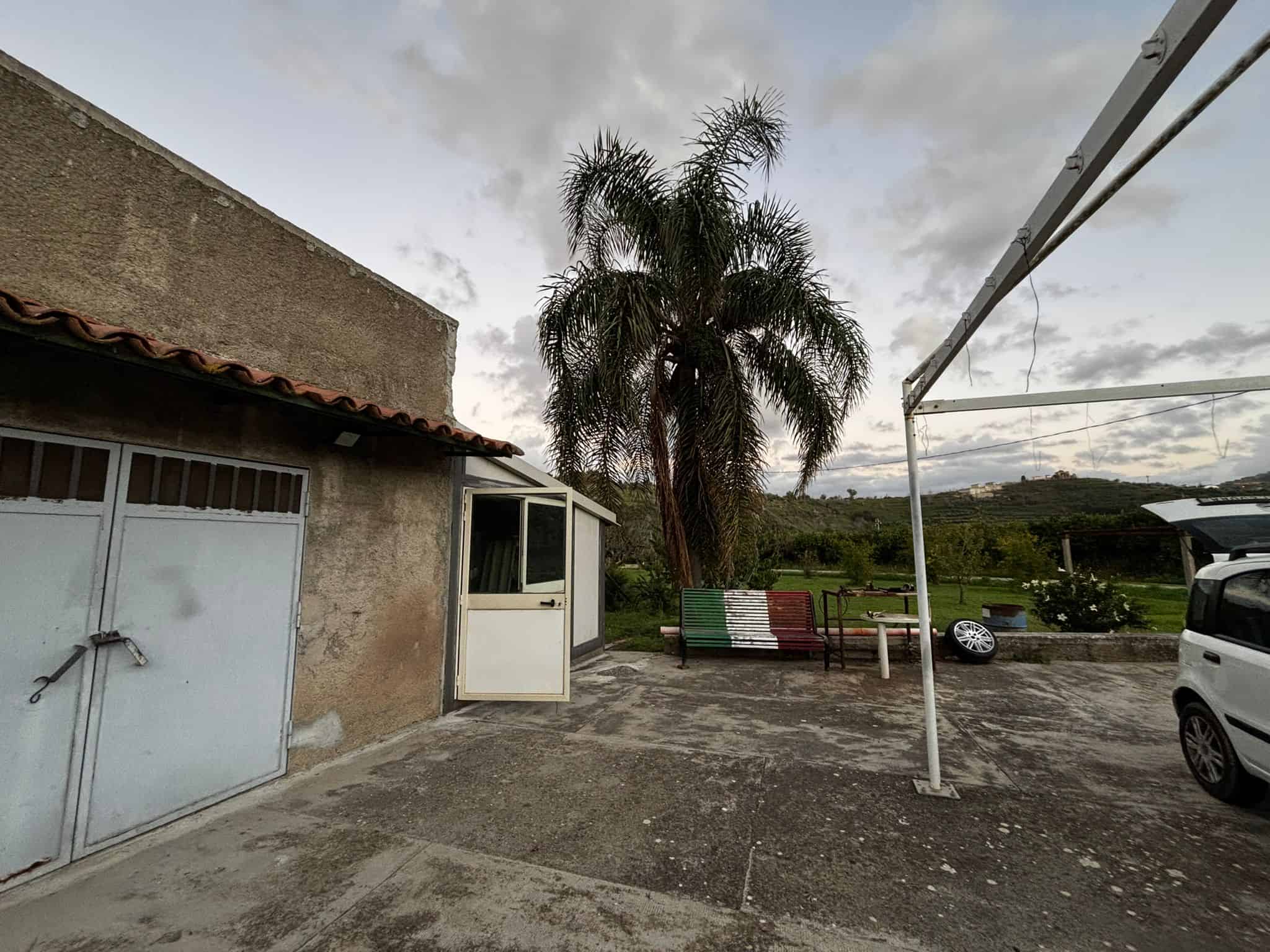 Tranquillo cortile esterno con alberi di palma, sedie colorate e auto, atmosfera rurale e rilassante, perfetto per godersi il paesaggio e il relax all'aperto in campagna.