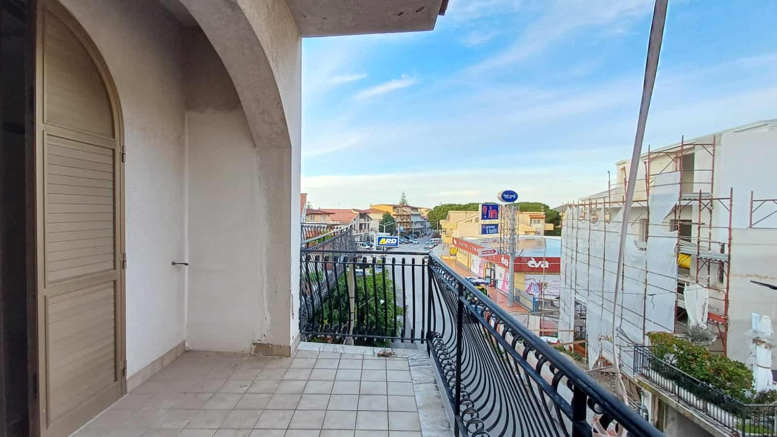 Vista panoramica di un balcone con ringhiera in ferro battuto, affaccio su via commerciale con negozi e segnaletica pubblicitaria, edificio in fase di ristrutturazione, cielo sereno, location urbana e di facile accesso.