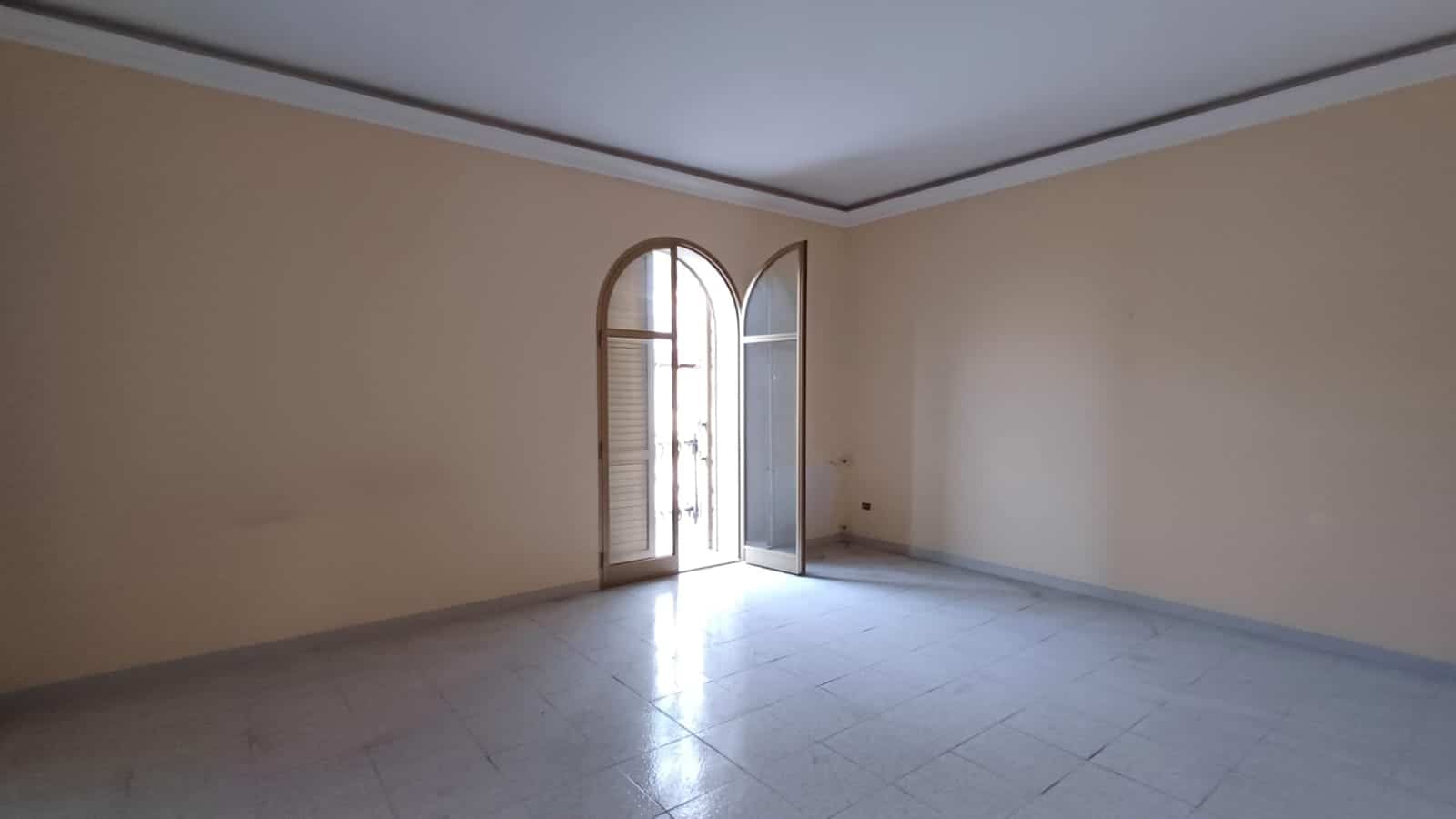 Ampia stanza luminosa in affitto, con pareti beige, grande finestra ad arco e porta finestra, perfetta per vivere in spazio e comfort. Ideale per residenze in zona tranquilla.