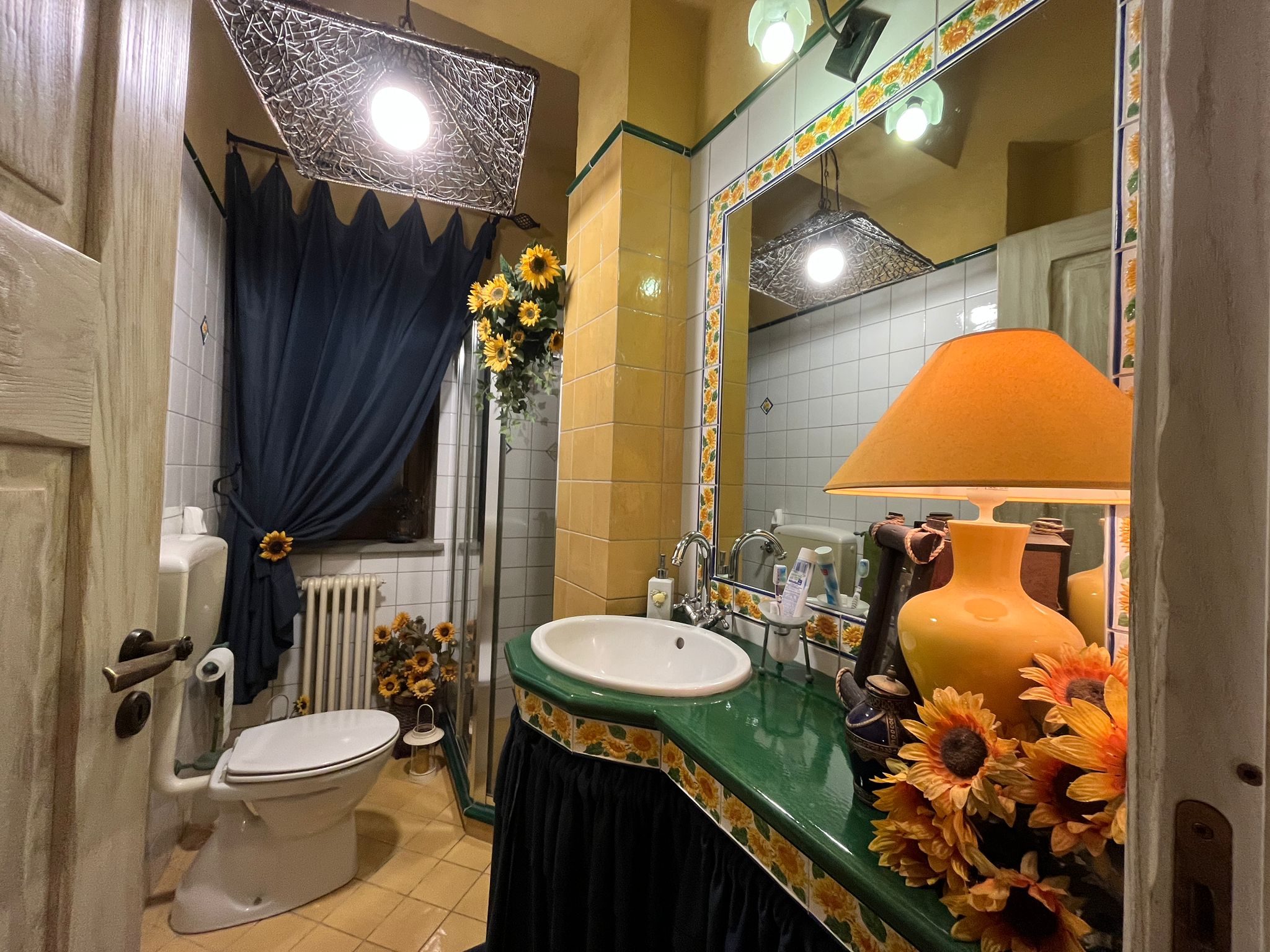 Lavabo con decoro floreale, specchio grande e dettagli di arredo con girasoli, bagno shabby chic con luci calde e dettagli vintage, ambiente accogliente e colorato.