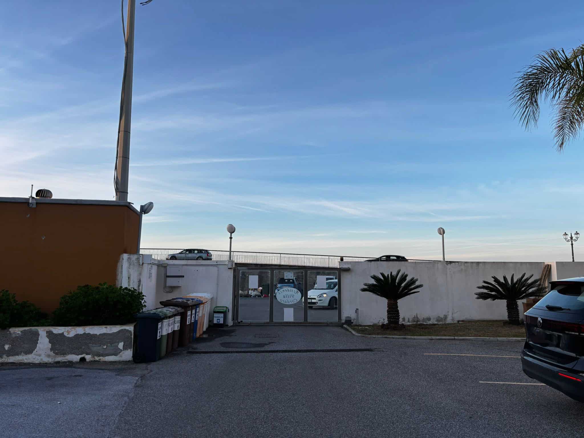 Prospettiva del parcheggio esterno di NextCasa con auto e albero di palma, atmosfera serale luminosa, ideale per servizi immobiliari e annunci di appartamenti.