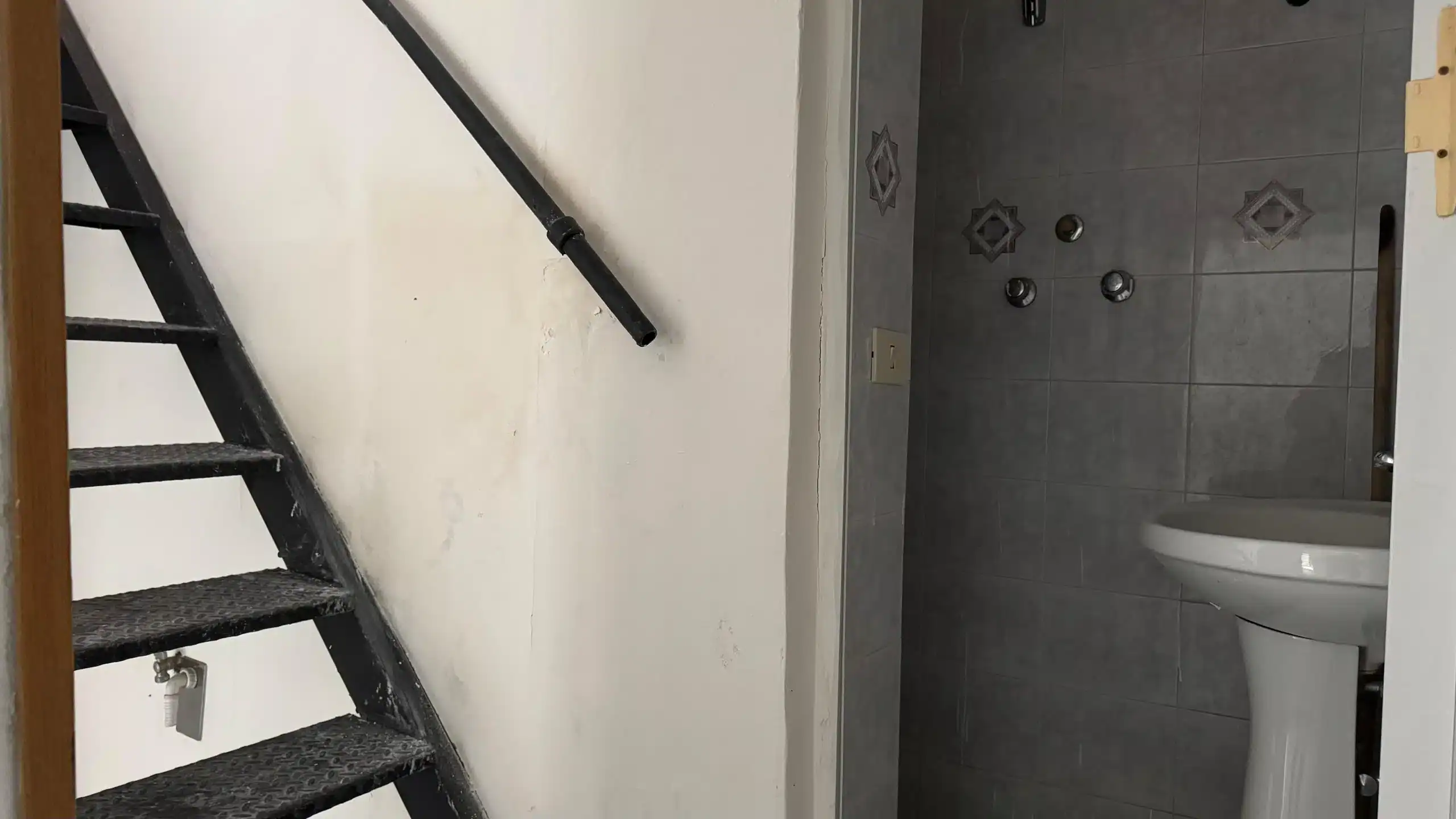Riduzione immobile in corso: vista delle scale nere e del bagno con piastrelle grigie, lavandino e bottoni dell'impianto idraulico internamente. Ideal for investitori o ristrutturatori.