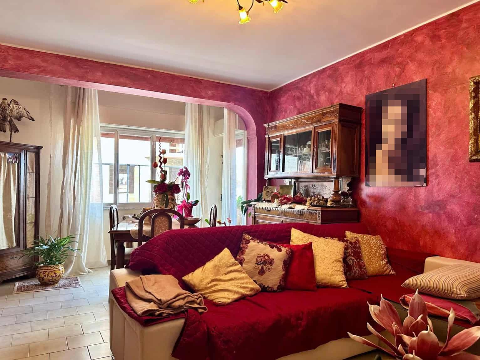 Colorata zona living con divano beige, cuscini decorativi multicolore, pareti rosso intenso, mobili in legno antico, dettaglio di decorazioni floreali e una finestra luminosa con tende chiare.