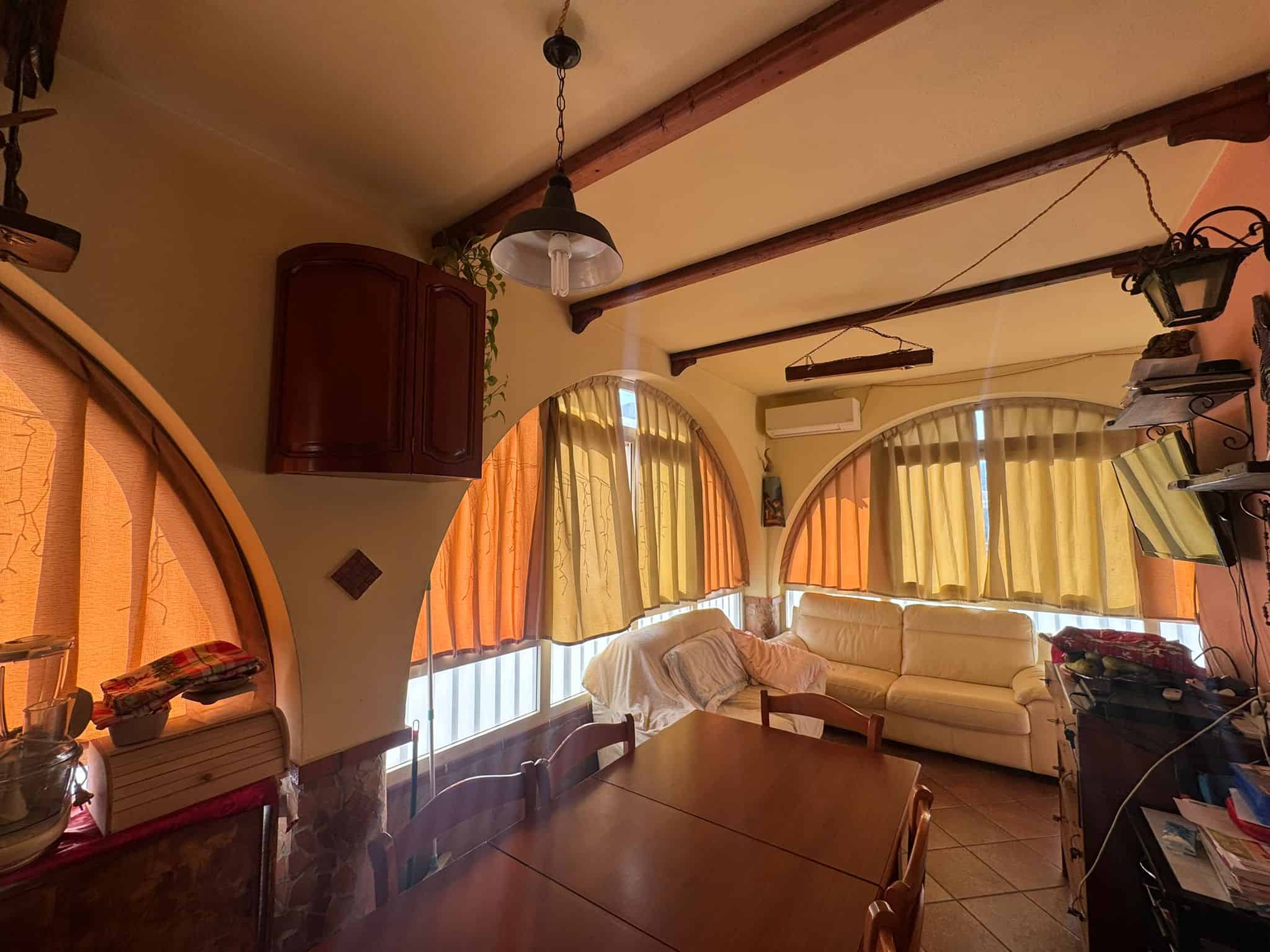 Soggiorno con grandi finestre, tende colorate, divano bianco e arredamenti in stile rustico, ideale per case o appartamenti da comprare o affittare, con comfort e carattere.