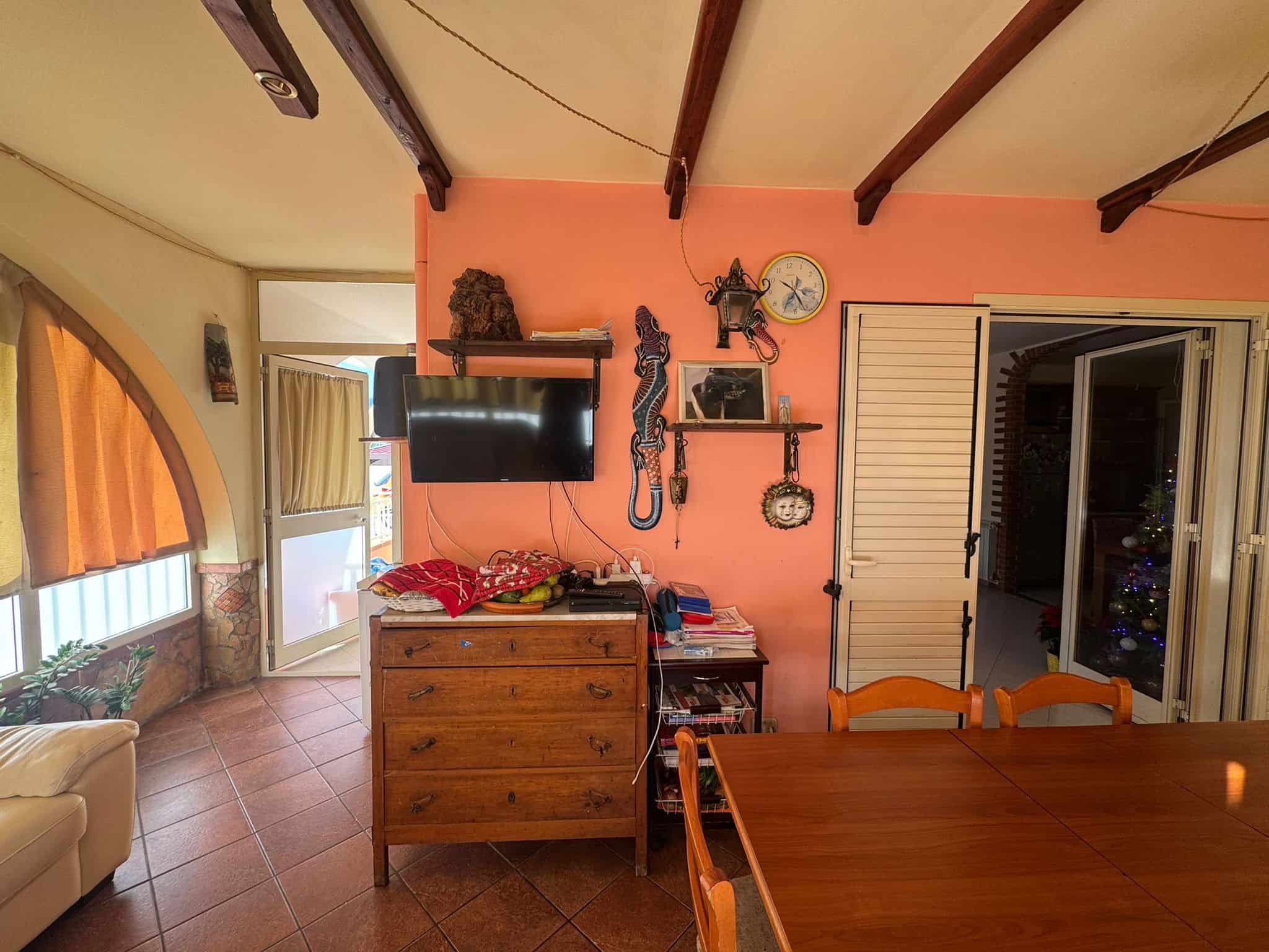 1. Interno soggiorno con pareti rosa, televisore, decorazioni artigianali e accessori, con ampie finestre e porta scorrevole che conduce a balcone, stile rustico e accogliente.