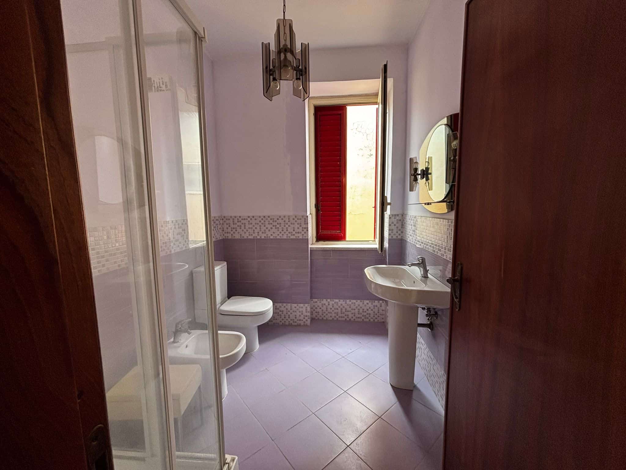 Bagno spazioso con lavabo, WC, bidet e cabina doccia, luce naturale dalla finestra con persiana rossa, piastrelle viola e mosaico decorativo, arredamento semplice, ideale per affitti e vendita immobili NextCasa.