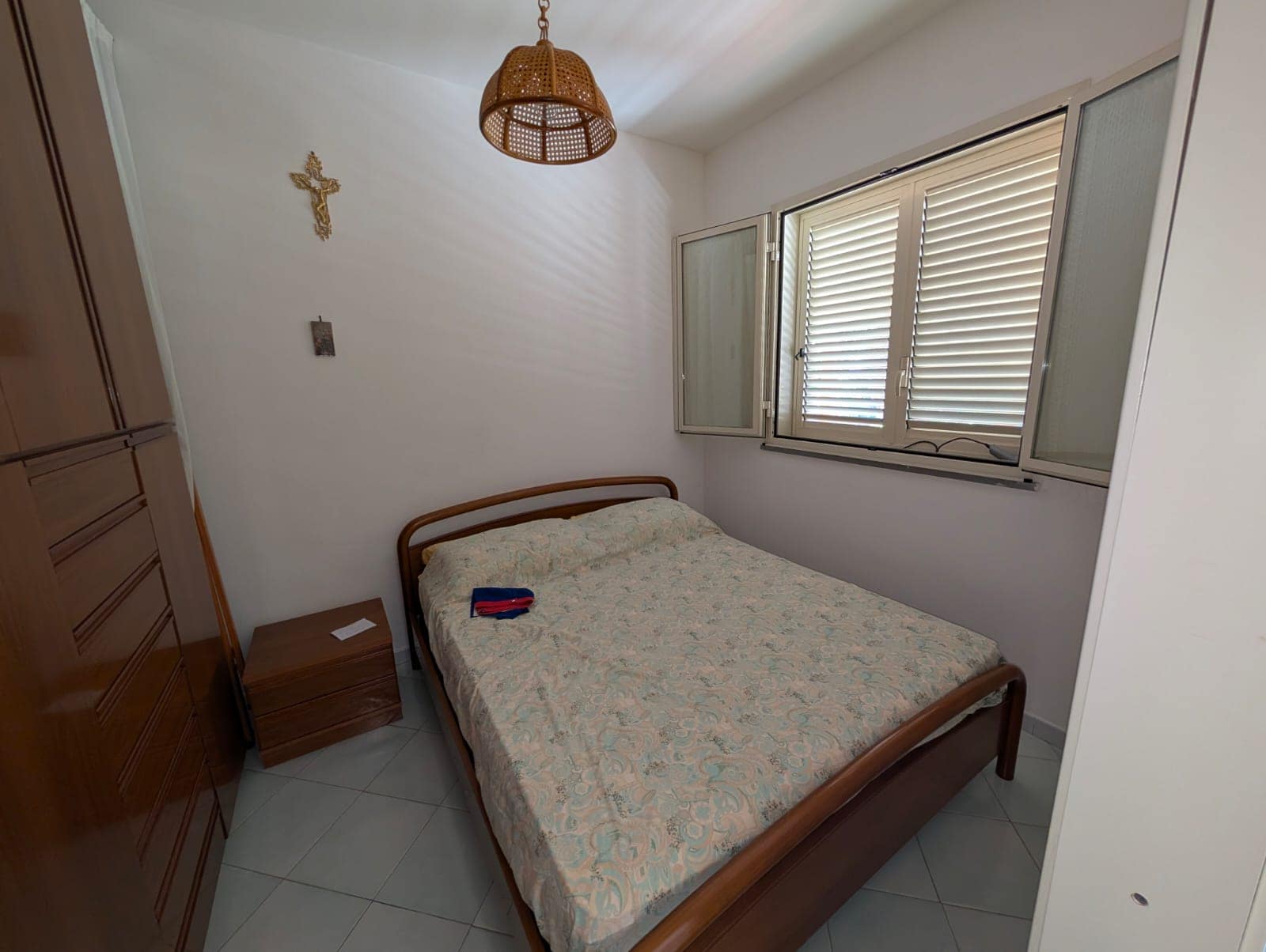 Letto matrimoniale in una stanza luminosa con finestra a battente e tende veneziane, arredi in legno chiaro, pareti bianche e decorazioni religiose, atmosfera semplice e accogliente.