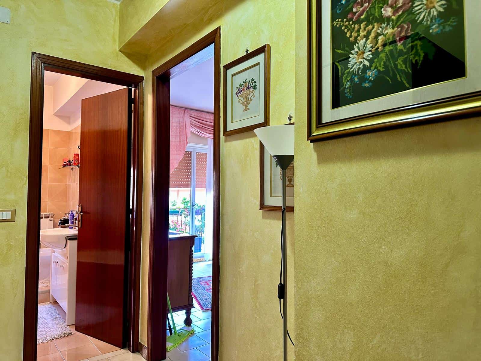 Ingresso di una casa con pareti color giallo, quadri floreali alle pareti, lampada da terra e vista su balcone e soggiorno con tende rosa e mobile in legno.