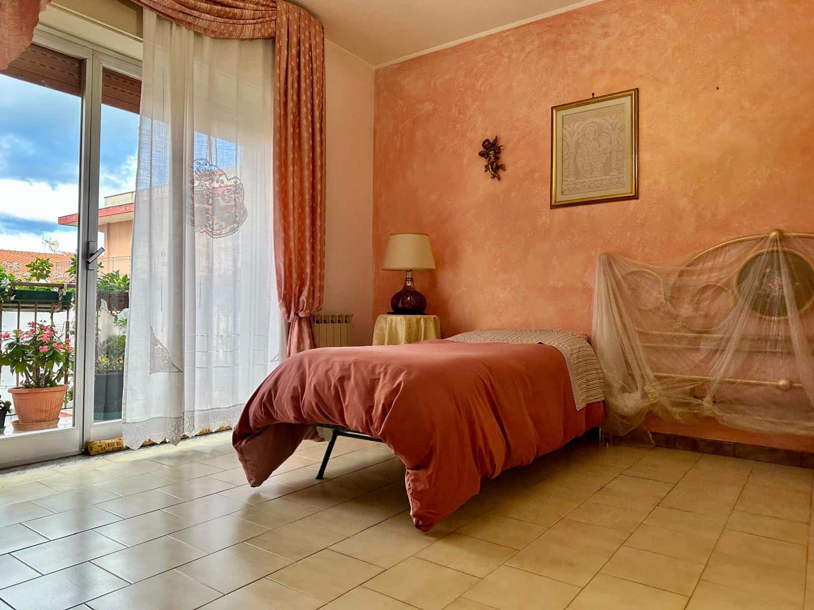 Arcobaleno di luce naturale sulla camera da letto con pareti rosee, tendaggi eleganti, balcone con piante, letto matrimoniale con zanzariera, ideale per immobili in vendita o affitto con servizi NextCasa.