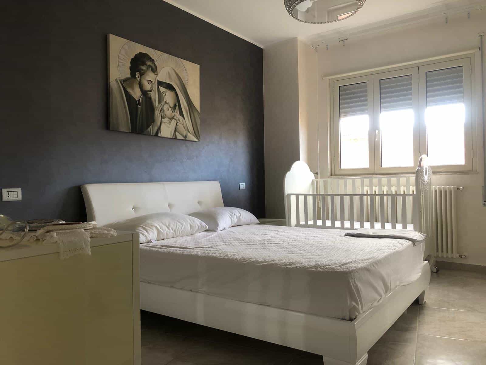 Camera da letto moderna con letto matrimoniale bianco, parete accentuata grigia, ampia finestra con vista luminosa, decorazione religiosa sopra il letto, stile elegante e accogliente, ideale per immobili di pregio.