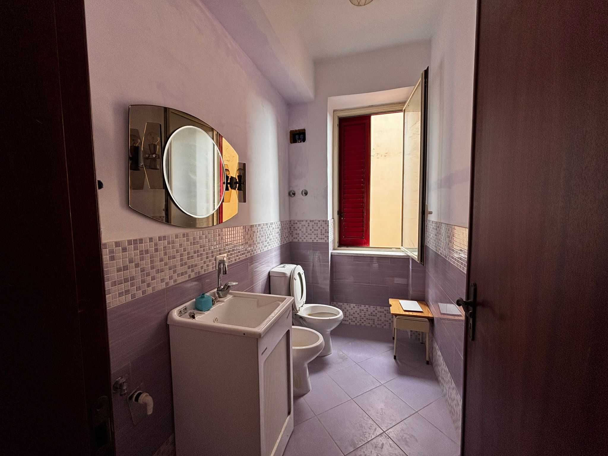 Lavanderia luminosa con finestra aperta, lavabo, toilette e piastrelle decorative in viola e beige, ideale per servizi domestici e organizzazione spazi in immobili a Roma.