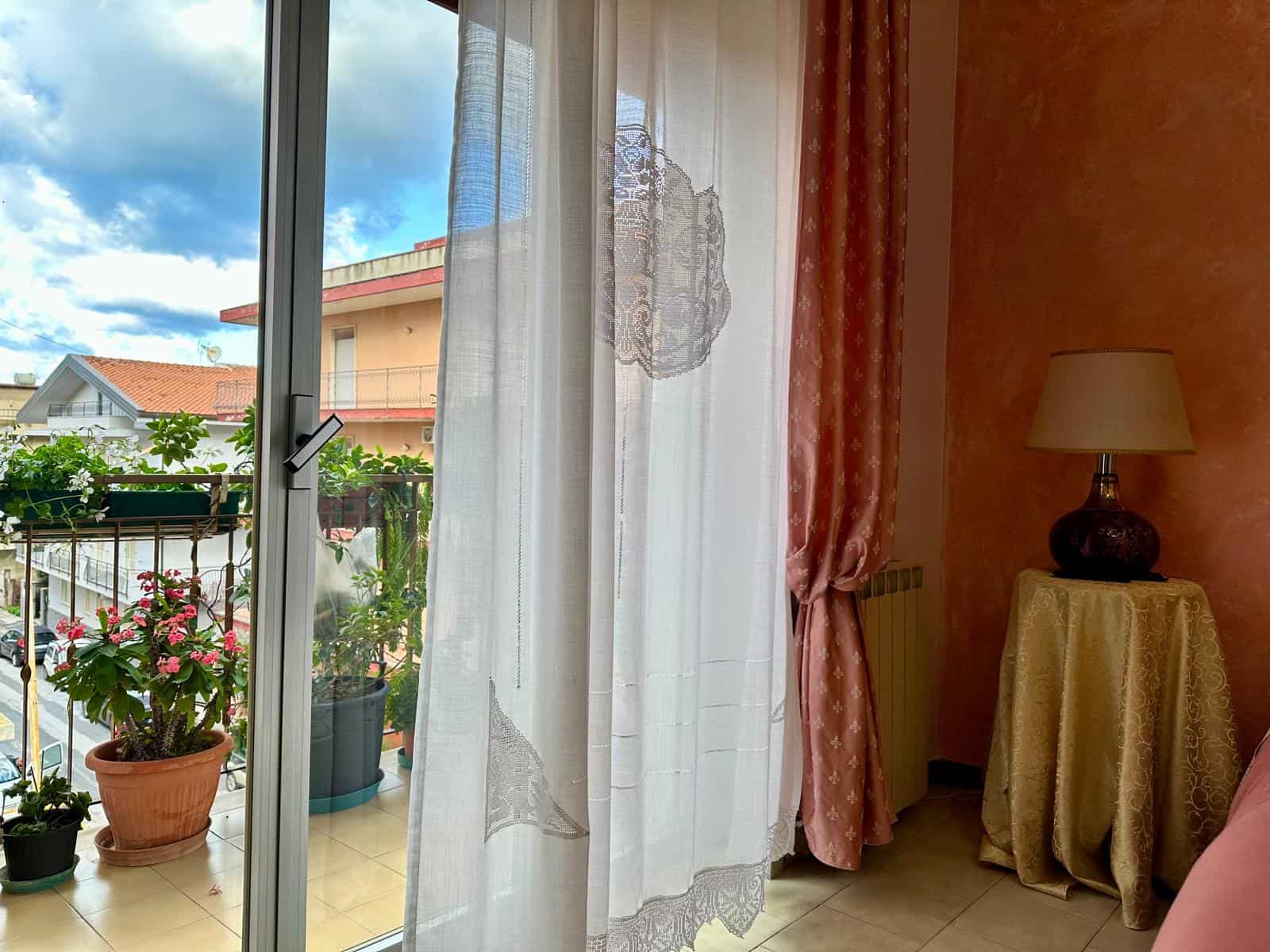 Balcone con piante fiorite, grande finestra con tende leggere, luce naturale che entra in stanza con pareti rosa e lampada decorativa.