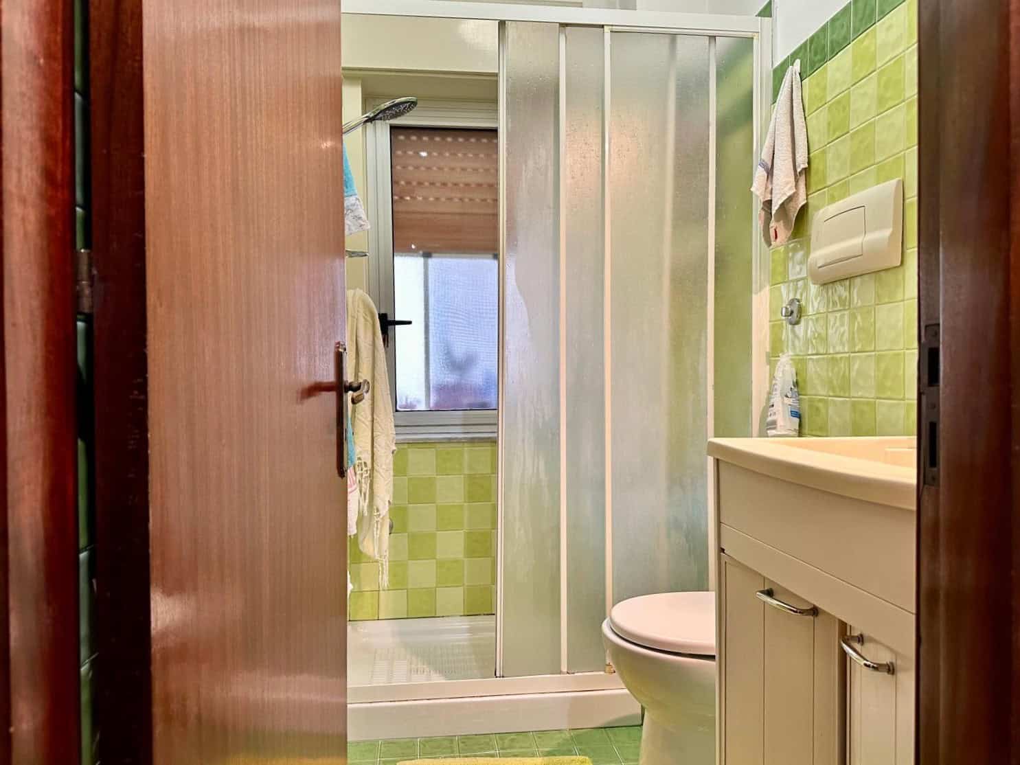 Bagno luminoso con piastrelle verdi, doccia, finestra, lavabo con mobiletto, elementi di arredo e accessori bagno, arredamento funzionale e colori vivaci per uno spazio bagno accogliente e pratico.