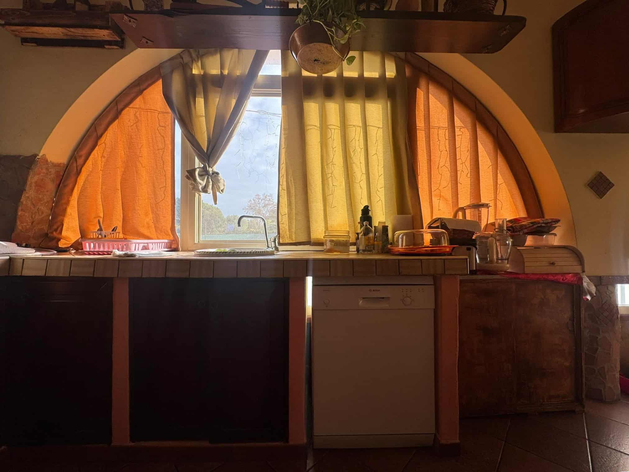 Pannello della cucina con tende arancioni e gialle sotto una grande arcata, vista verso l'esterno con paesaggio naturale, luce naturale, ambiente accogliente e rustico.